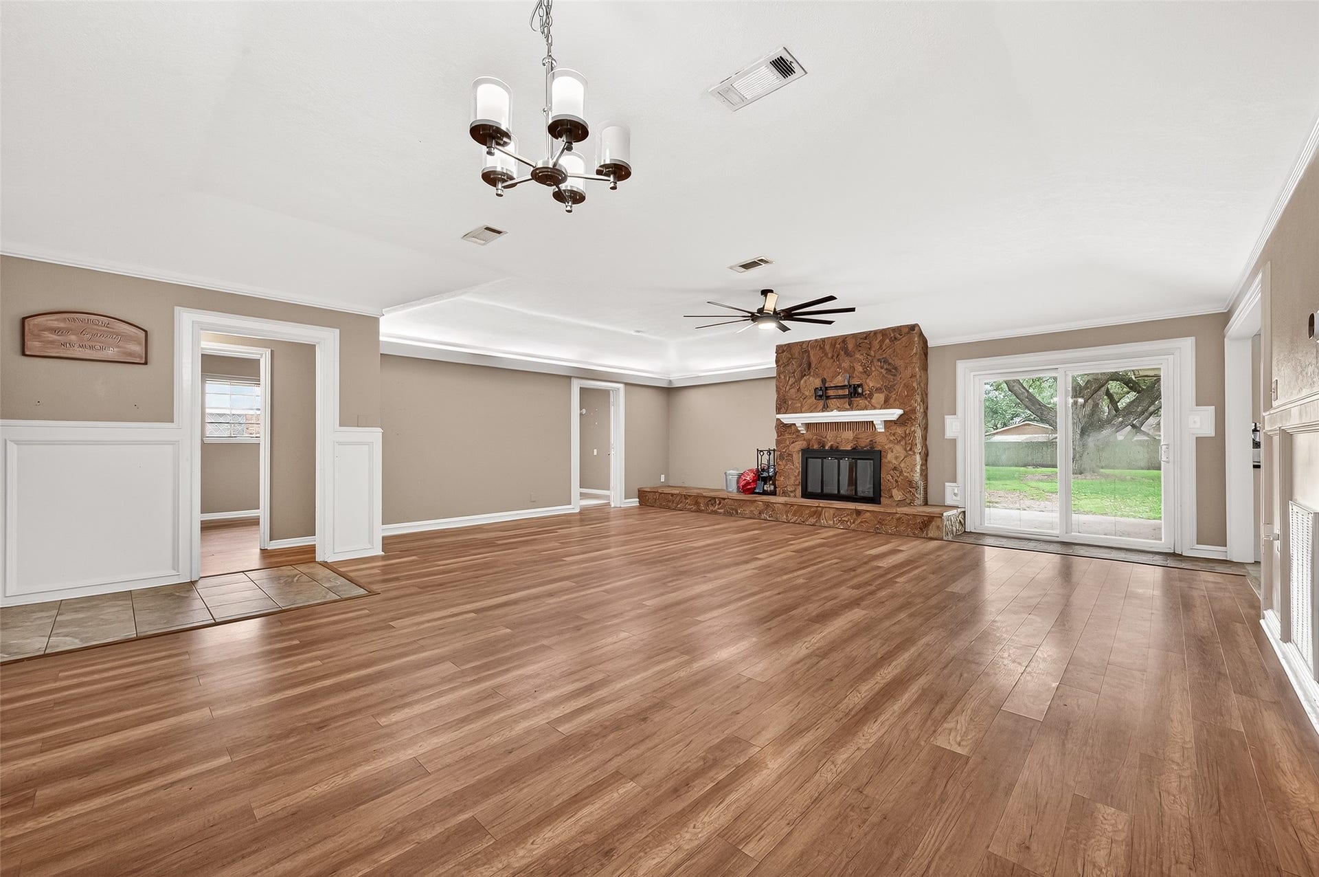 5607 Avenue M 1/2, Santa Fe, TX 77510 Spacious living room with elegant wood flooring and a cozy stone fireplace. Features a modern ceiling fan and a classic chandelier. Large glass doors offer a scenic view and natural light, leading to the backyard. Neutral wall tones provide a warm, inviting atmosphere.