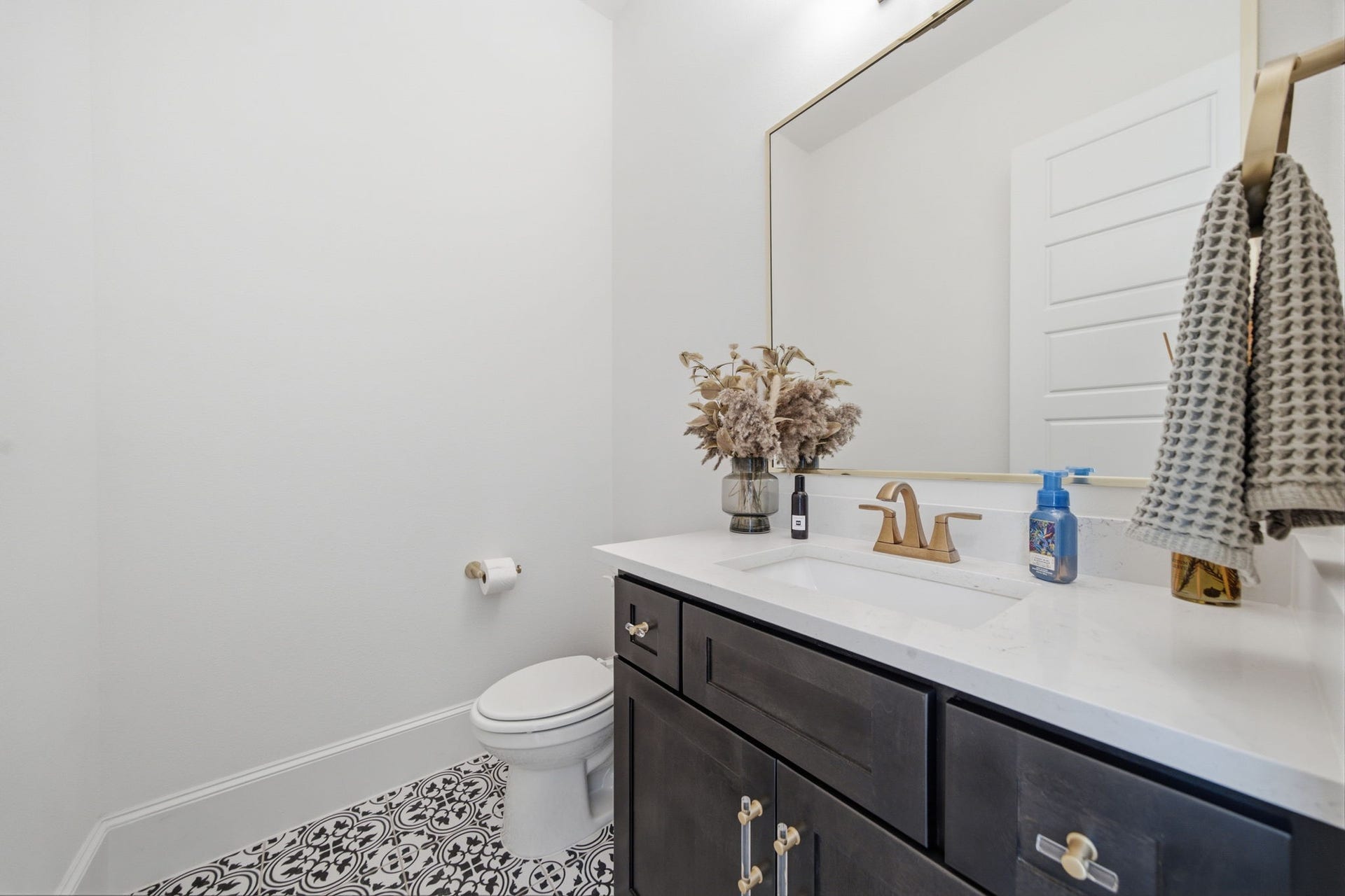 6310 Star Light Court, Spring, TX 77386 Elegant powder room featuring a sleek vanity with a quartz countertop, accented by a large mirror and stylish brass fixtures. The patterned tile floor adds a touch of sophistication.