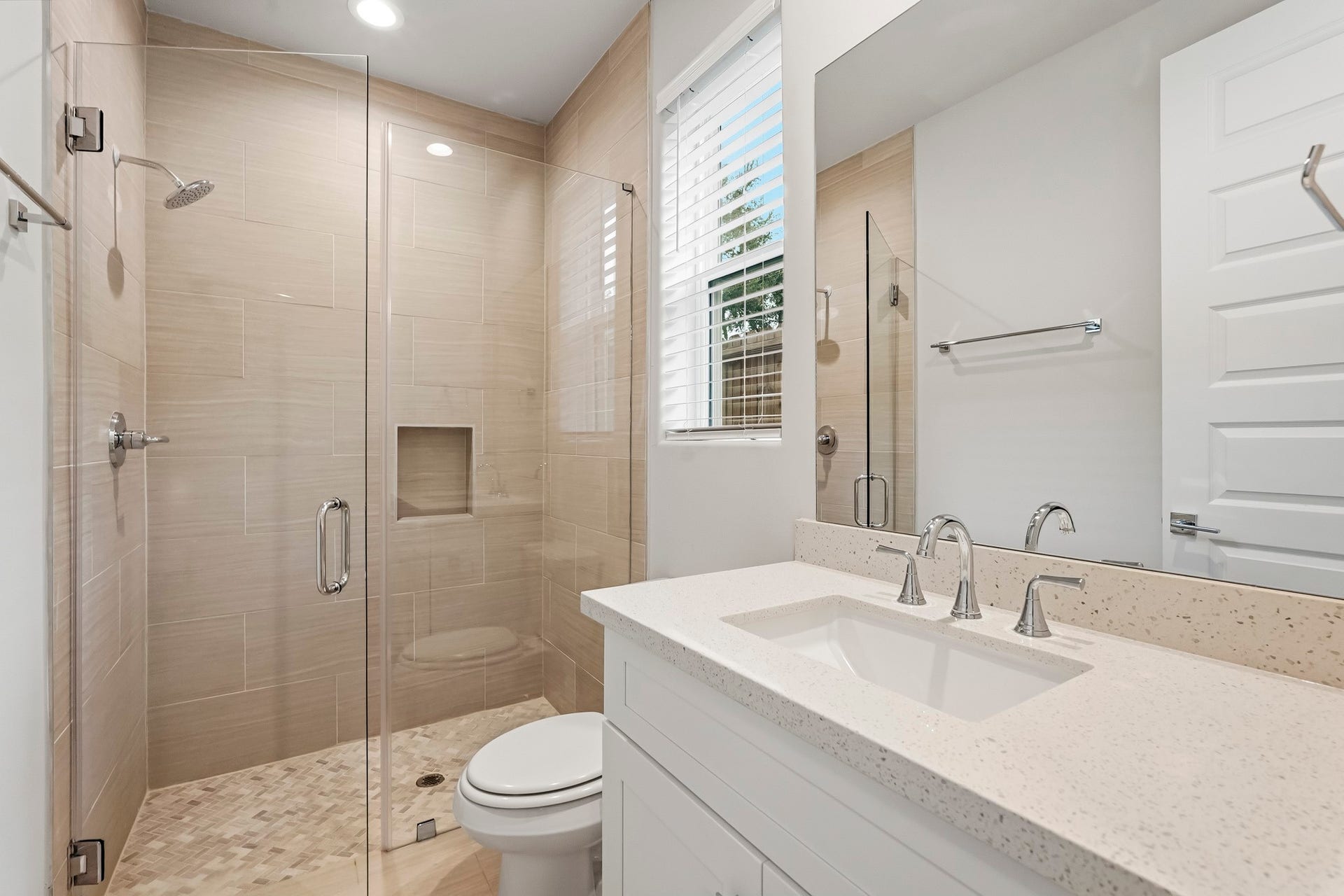 3109 Arlington Street, Houston, TX 77018 The bright and modern en-suite bathroom features a spacious walk-in shower with frameless glass enclosure, neutral tile surround, mosaic shower flooring, rainfall-style showerhead, and a built-in wall niche. The white vanity is topped with a light quartz countertop, undermount sink, polished chrome fixtures, and a large wall mirror. A window with faux-wood blinds brings in natural light, while recessed lighting and clean, neutral finishes create a fresh and inviting atmosphere.