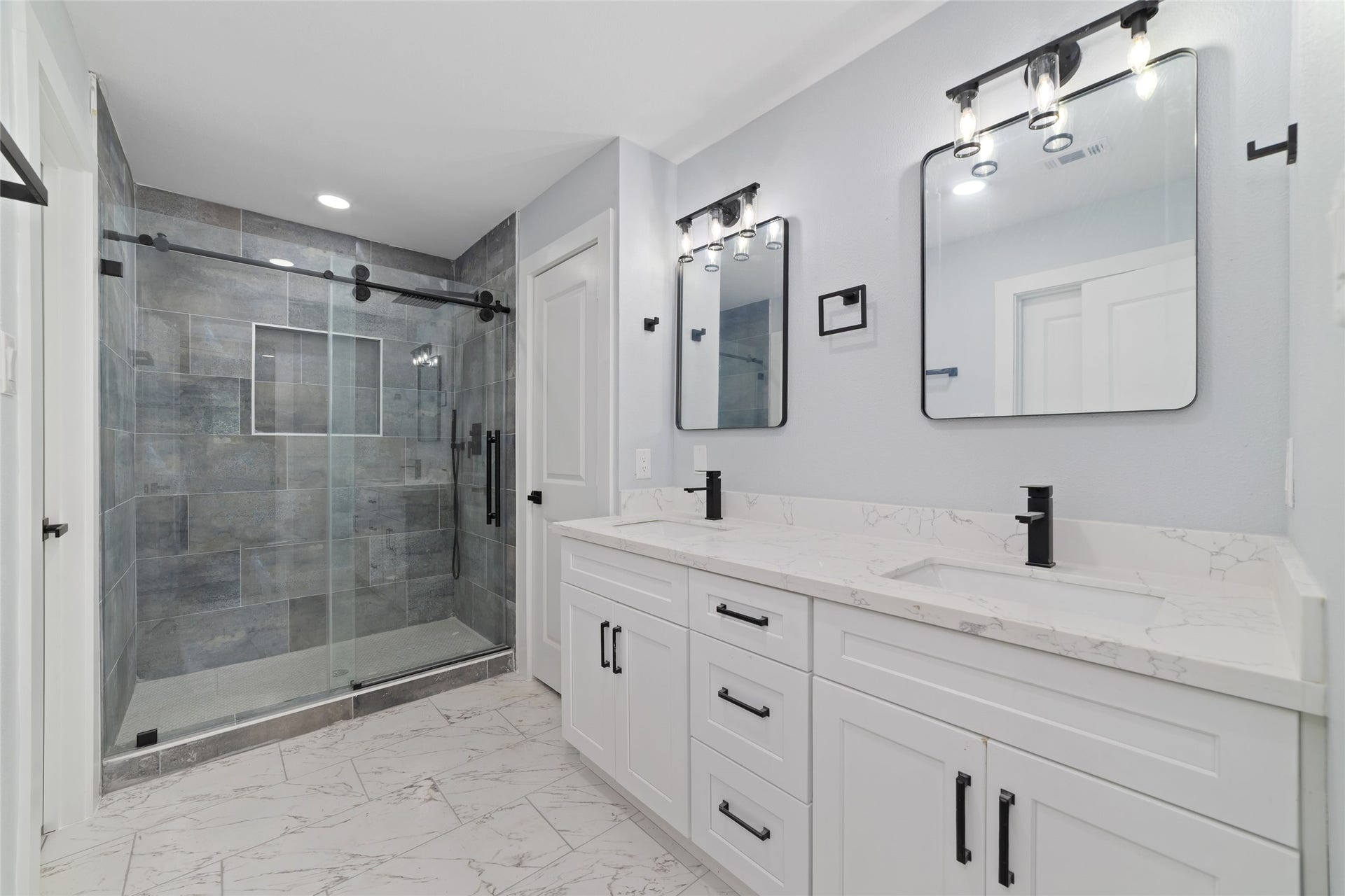A double-sink vanity, individual mirrors, and contrasting black fixtures complement the glass-enclosed shower with gray tile and a recessed storage niche.
