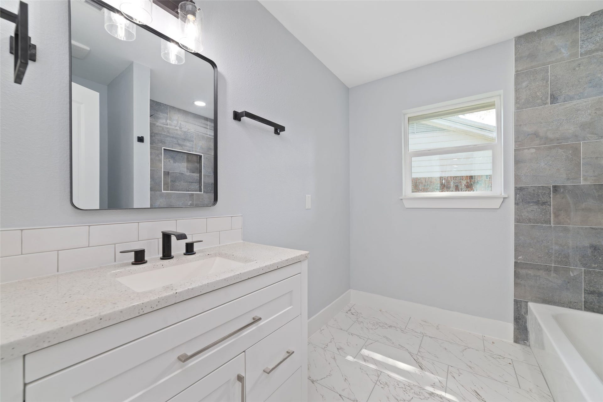 A light-colored vanity and black fixtures coordinate with the gray tile tub-and-shower surround. A recessed shower niche adds convenient storage.
