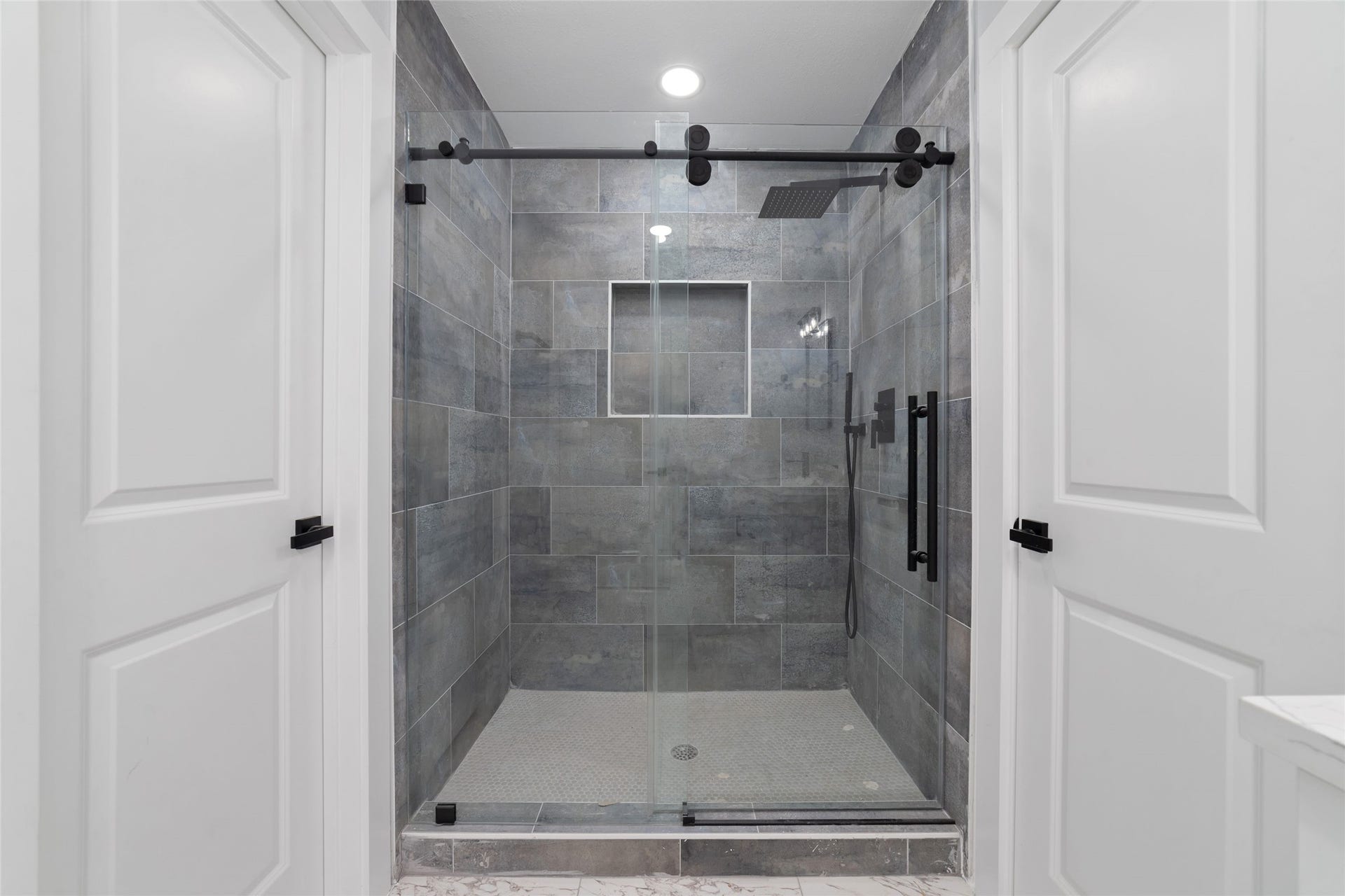 A double-sink vanity, individual mirrors, and contrasting black fixtures complement the glass-enclosed shower with gray tile and a recessed storage niche.