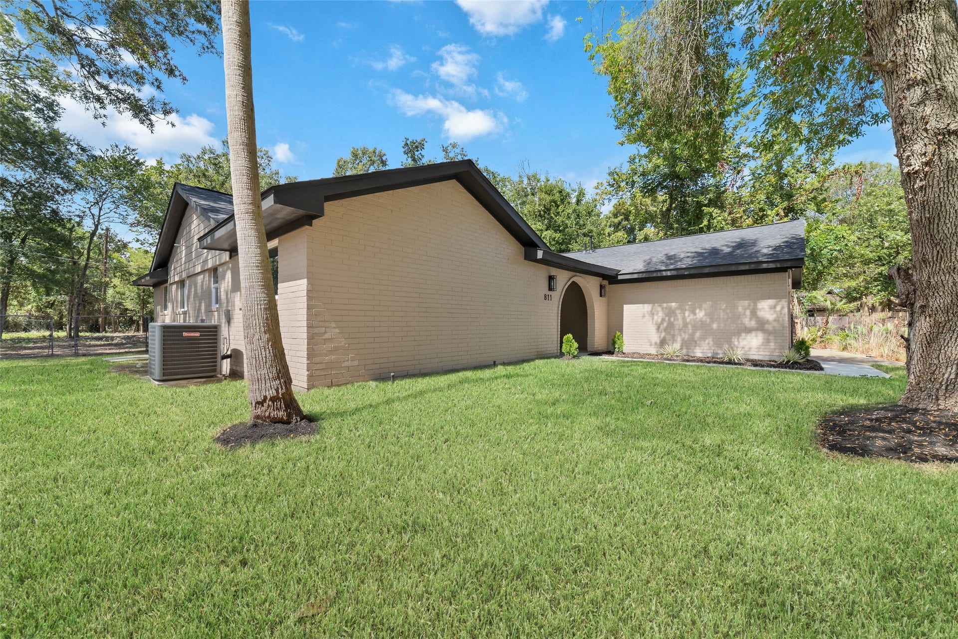Mature trees frame the front lawn, with a concrete walkway leading to the covered arched entrance.