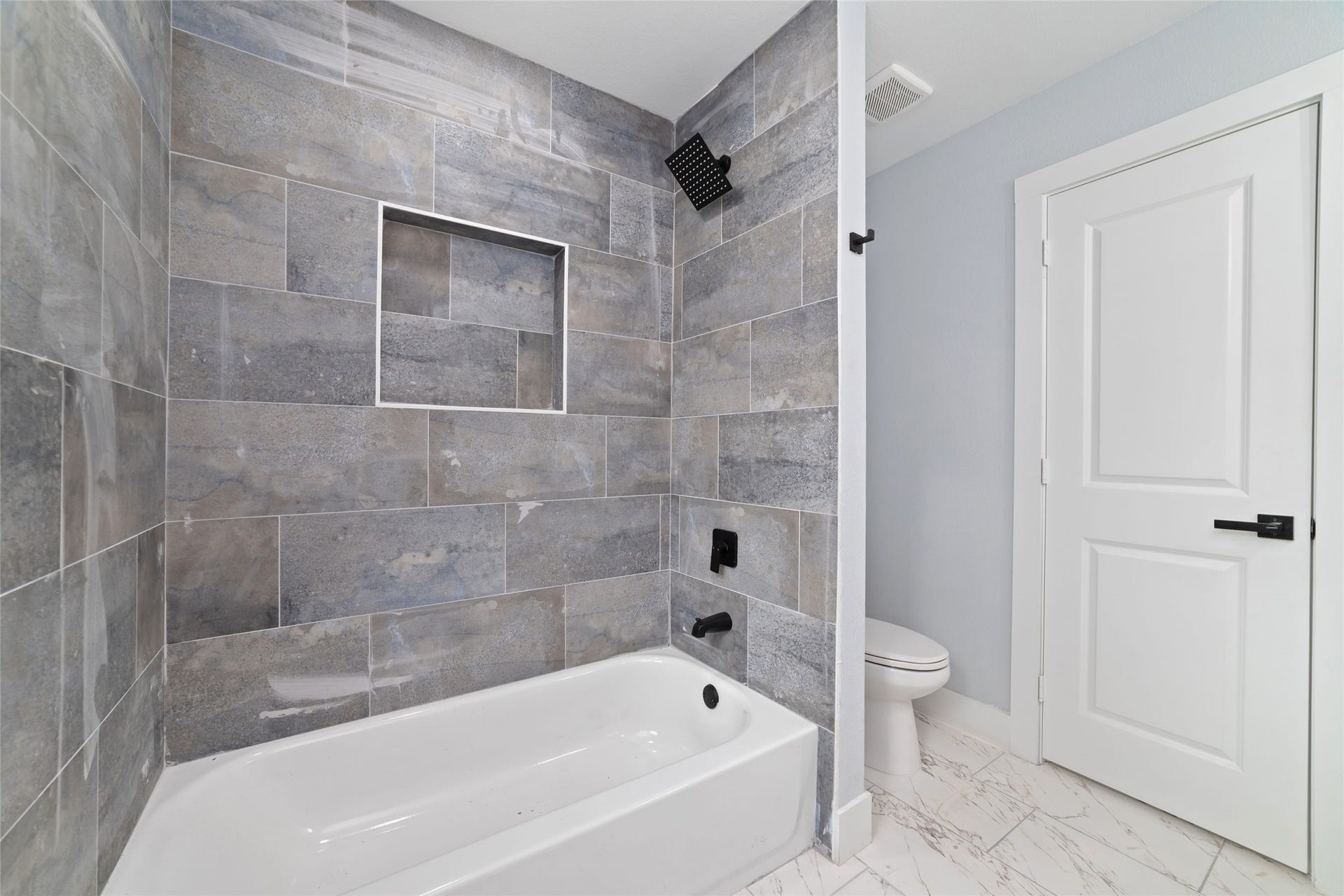 A light-colored vanity and black fixtures coordinate with the gray tile tub-and-shower surround. A recessed shower niche adds convenient storage.
