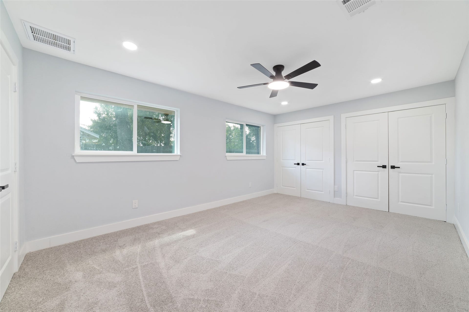 Multiple windows brighten this bedroom, which includes carpet flooring, a ceiling fan, and two double-door closets for storage.
