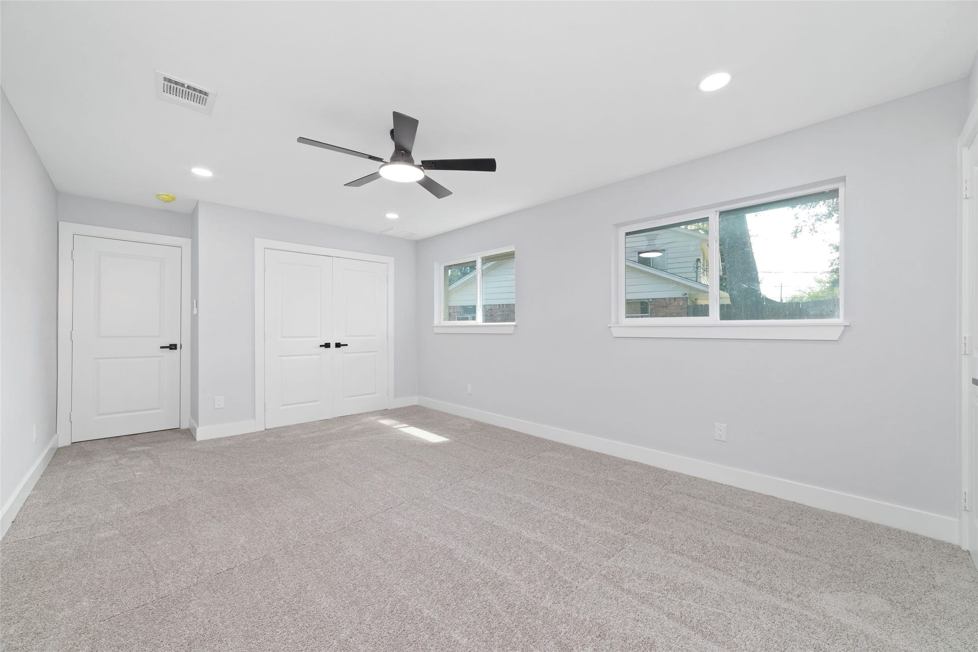 Multiple windows brighten this bedroom, which includes carpet flooring, a ceiling fan, and two double-door closets for storage.