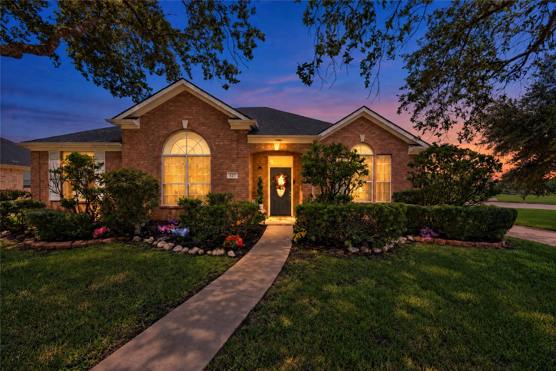 Single-story brick exterior at twilight featuring a arched entryway, large grand arched transom window, shuttered windows, warm interior and exterior accent lighting, concrete walkway, and landscaped garden beds with stone borders.