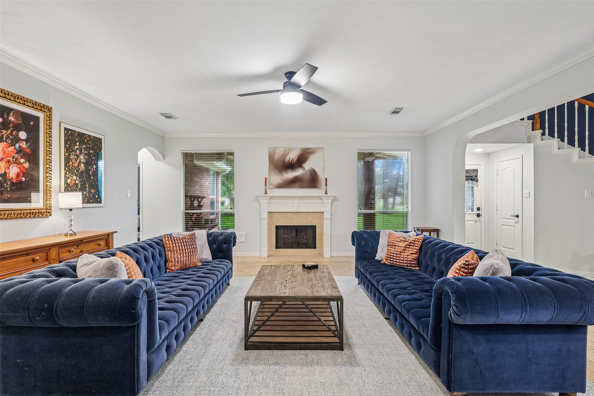 This inviting living room features two plush blue sofas, a central wooden coffee table, and a cozy fireplace. Large windows provide natural light, while tasteful artwork and a ceiling fan enhance the space. The open layout leads to a staircase and an adjoining room, creating a seamless flow.