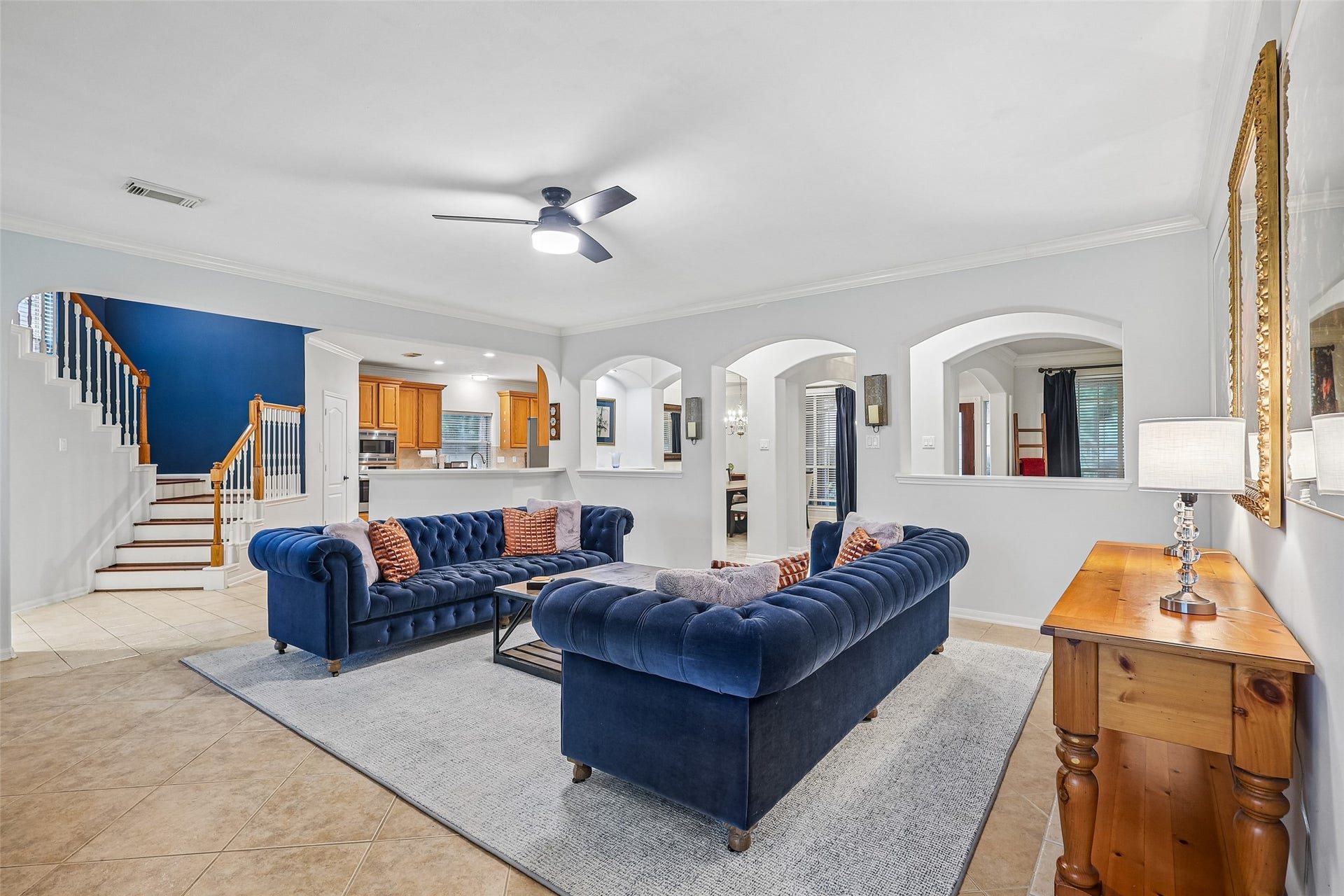 This inviting living room features elegant navy sofas, a modern ceiling fan, and a cozy atmosphere. The open layout connects seamlessly to a spacious kitchen with warm wooden cabinetry, while arched doorways add a touch of sophistication. A stairway with white and wooden accents leads to the upper level, enhancing the home's charm.