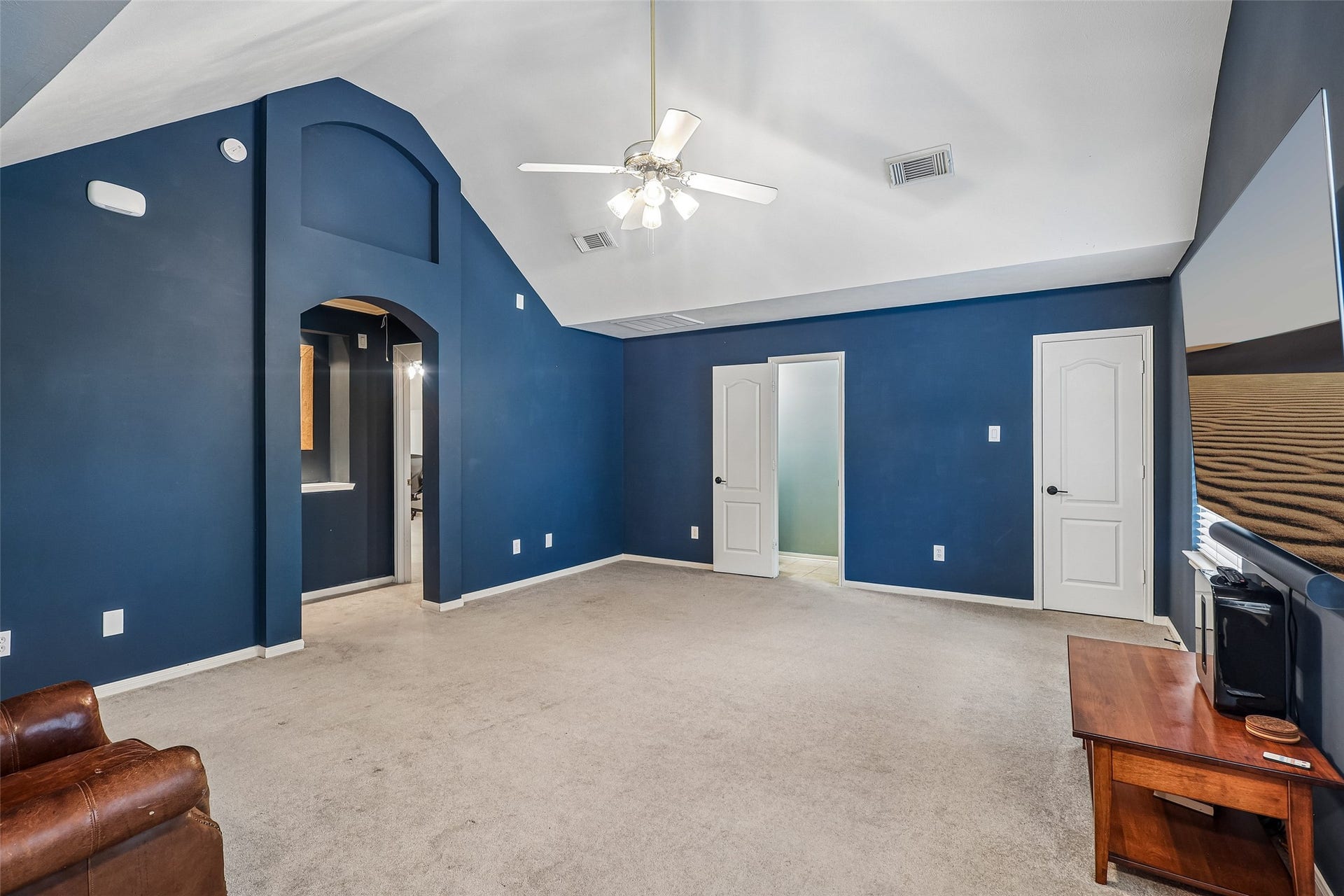 This spacious room features a high vaulted ceiling with a ceiling fan, dark blue walls, and neutral carpeting. It includes two white doors and an arched entryway, offering a versatile area for relaxation or entertainment.
