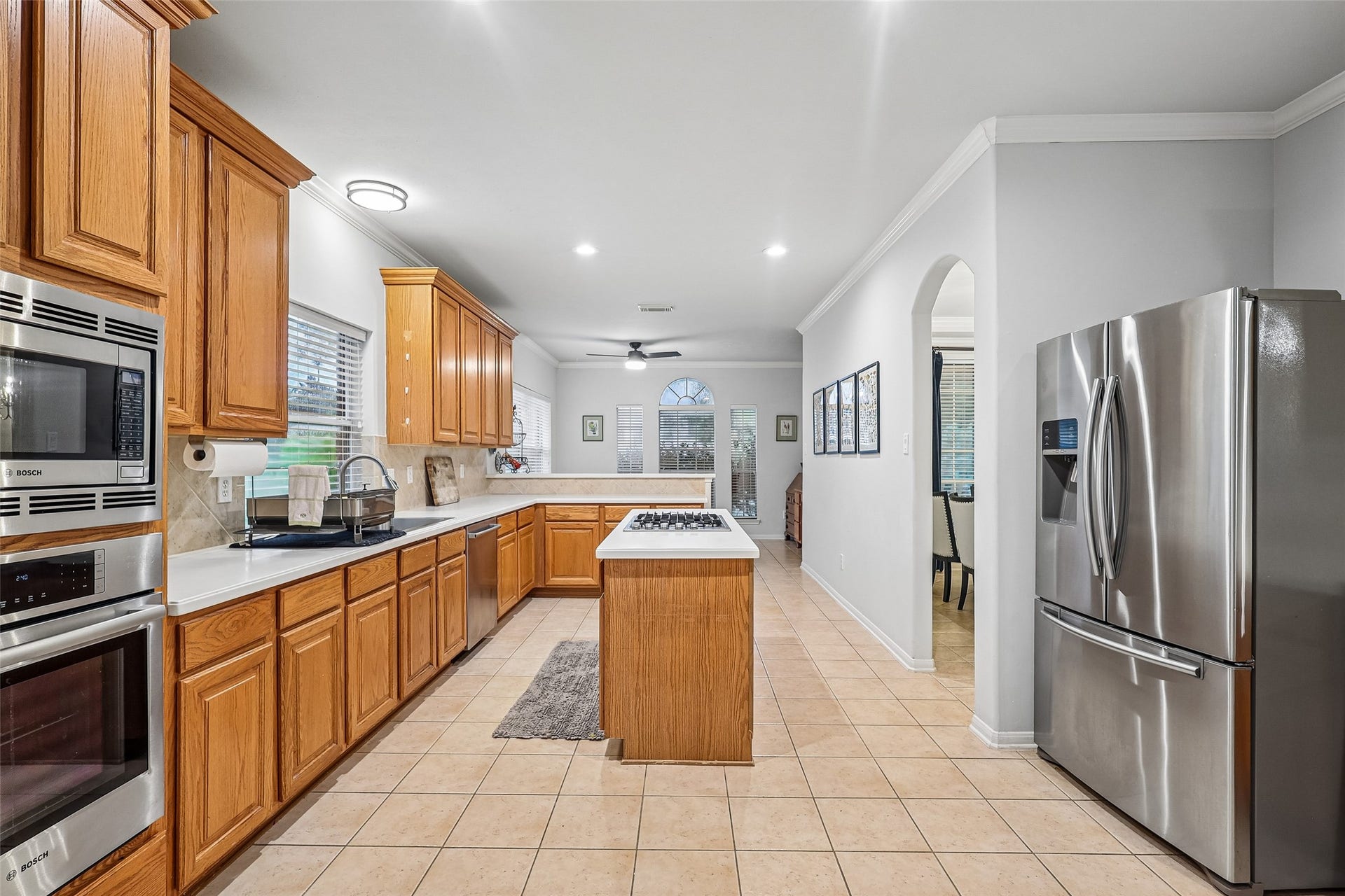 This modern kitchen features stainless steel appliances, ample wooden cabinetry, a central island with a cooktop, and a sleek, spacious layout with tile flooring and neutral walls, offering a bright and functional space for cooking and entertaining.