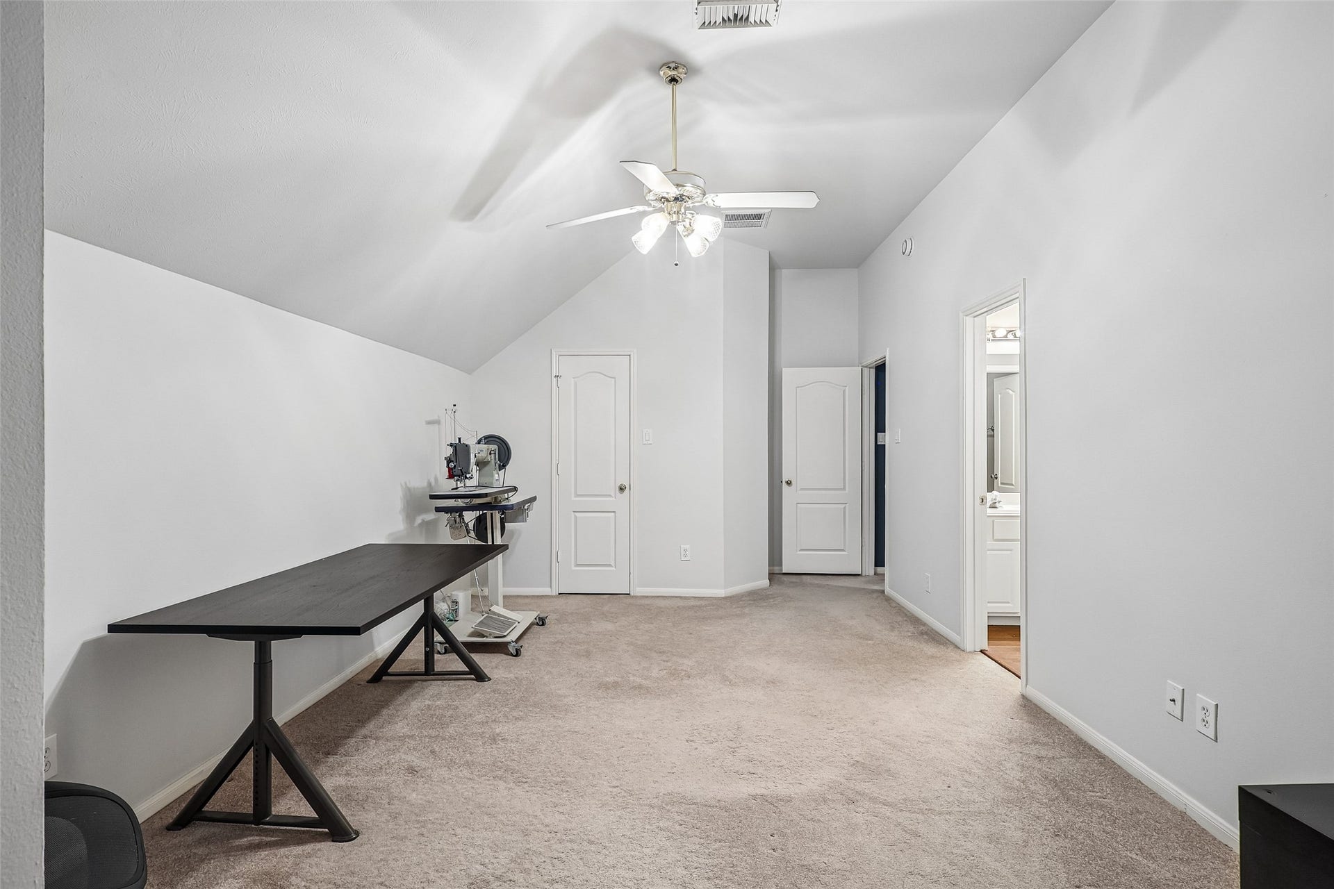 This room features a sloped ceiling with a ceiling fan and neutral carpet, offering a spacious area with a dark wood table. It includes access to an adjoining room or bathroom, making it versatile for office or hobby use.
