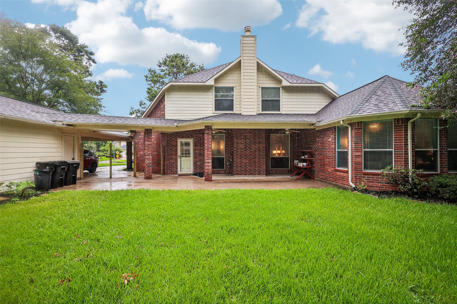 This photo shows a charming two-story brick home with a spacious, well-maintained backyard. It features a covered patio area and a detached garage connected by a carport. The exterior is accented with large windows and a chimney, offering a cozy and inviting atmosphere.
