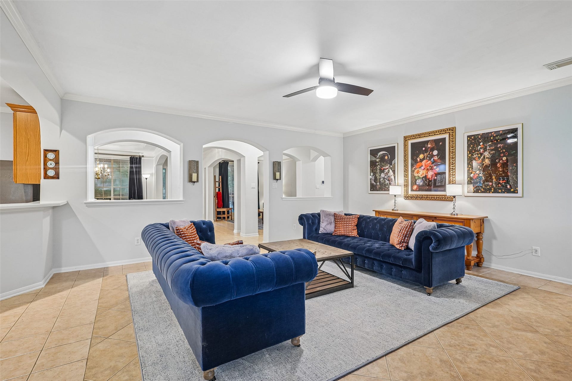 Spacious living room with elegant blue sofas and modern decor. Features tiled flooring, a ceiling fan, and stylish wall art. Arched doorways create an open, inviting atmosphere with plenty of natural light.