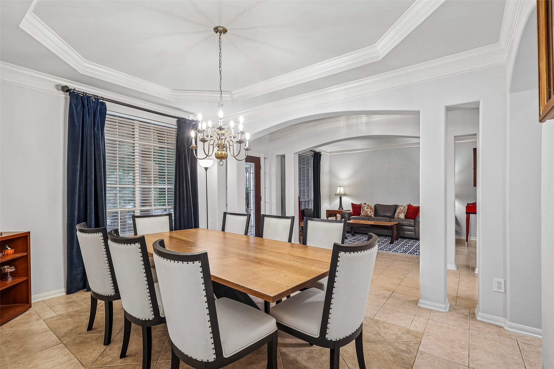 This inviting dining area features a spacious wooden table with elegant chairs, a stylish chandelier, and large windows with dark curtains. It opens into a cozy living space with comfortable seating and tasteful decor.