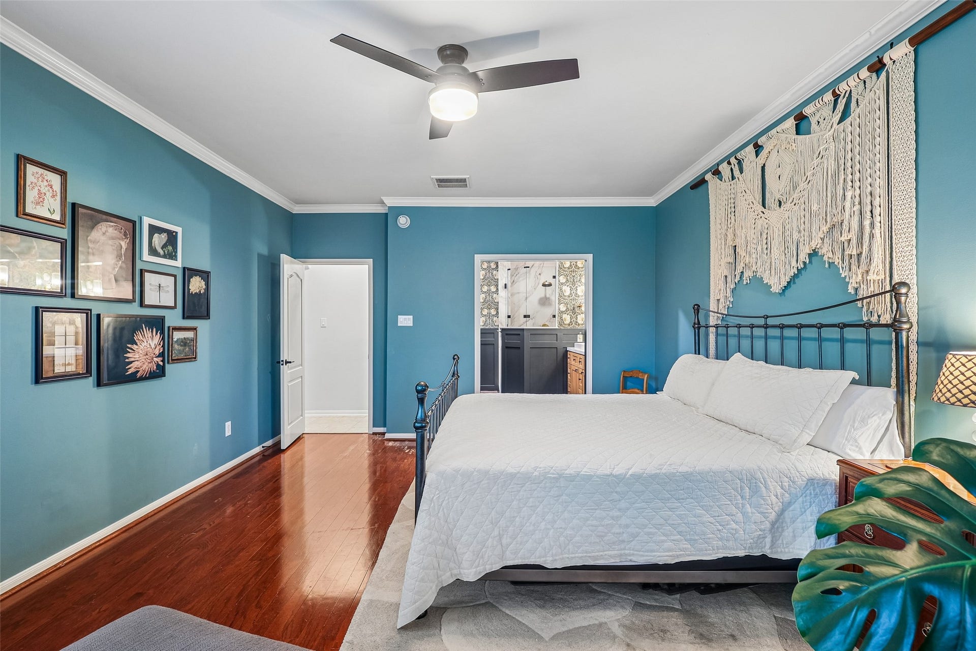 This bedroom features a soothing blue color scheme with a cozy feel, highlighted by a decorative macram wall hanging above a metal-framed bed. The room includes a ceiling fan, polished wood floors, and is accented with framed artwork. An en-suite bathroom is visible through an open door, providing convenience and style.