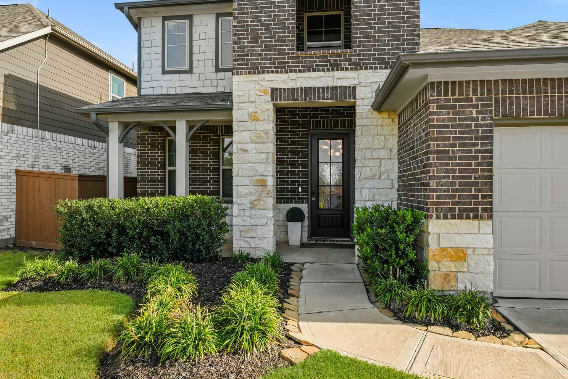 This inviting front entry showcases a handsome blend of brick and stone, complemented by manicured landscaping and a covered porch. A curved walkway leads to the glass-paneled front door, while mature shrubs and neatly maintained garden beds enhance the homes welcoming curb appeal.