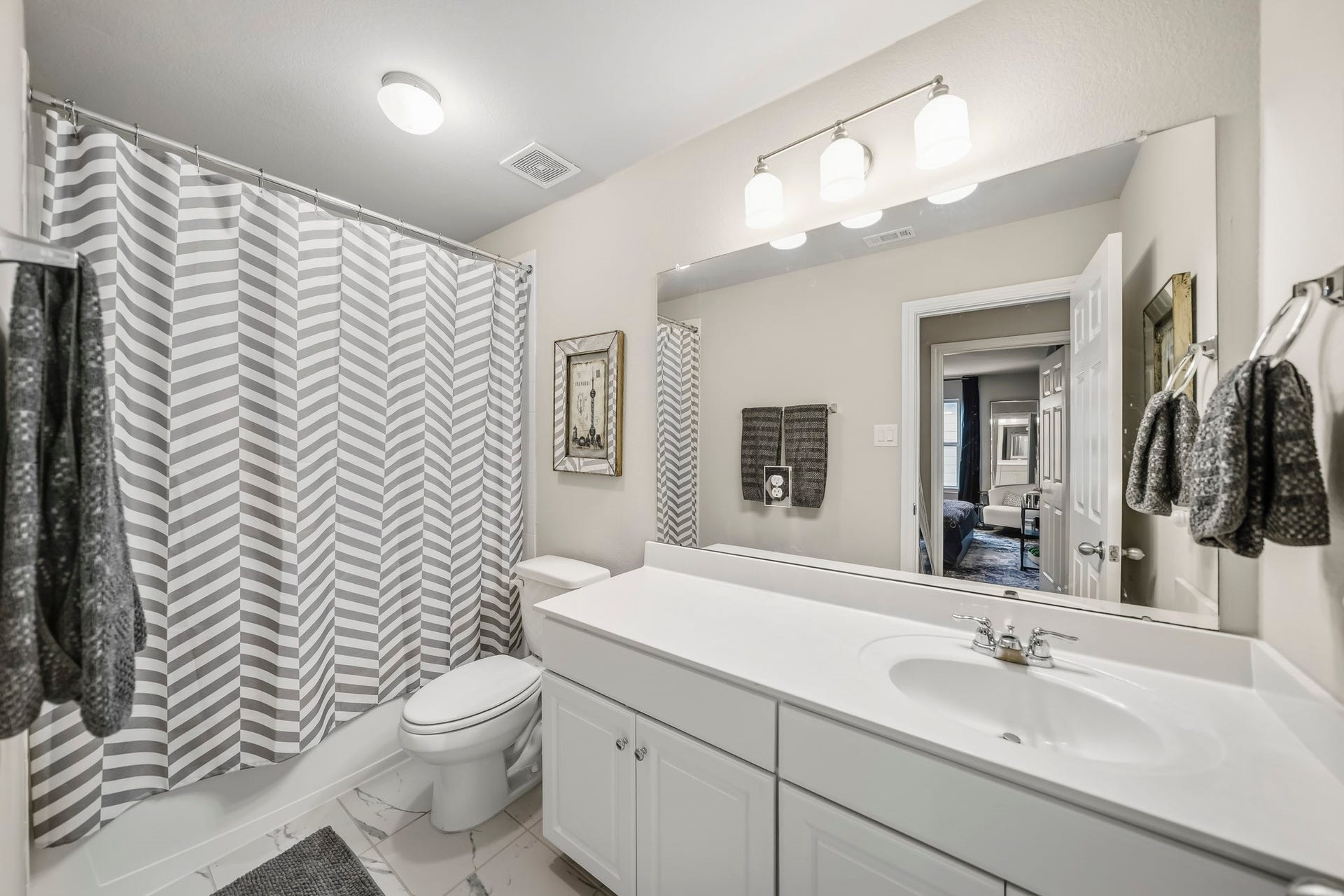 This bright secondary bathroom features a clean, neutral design with a spacious vanity, generous countertop space, and a large wall-to-wall mirror. A tub and shower combination adds everyday functionality, while updated tile flooring and ample lighting create a fresh, inviting feel.