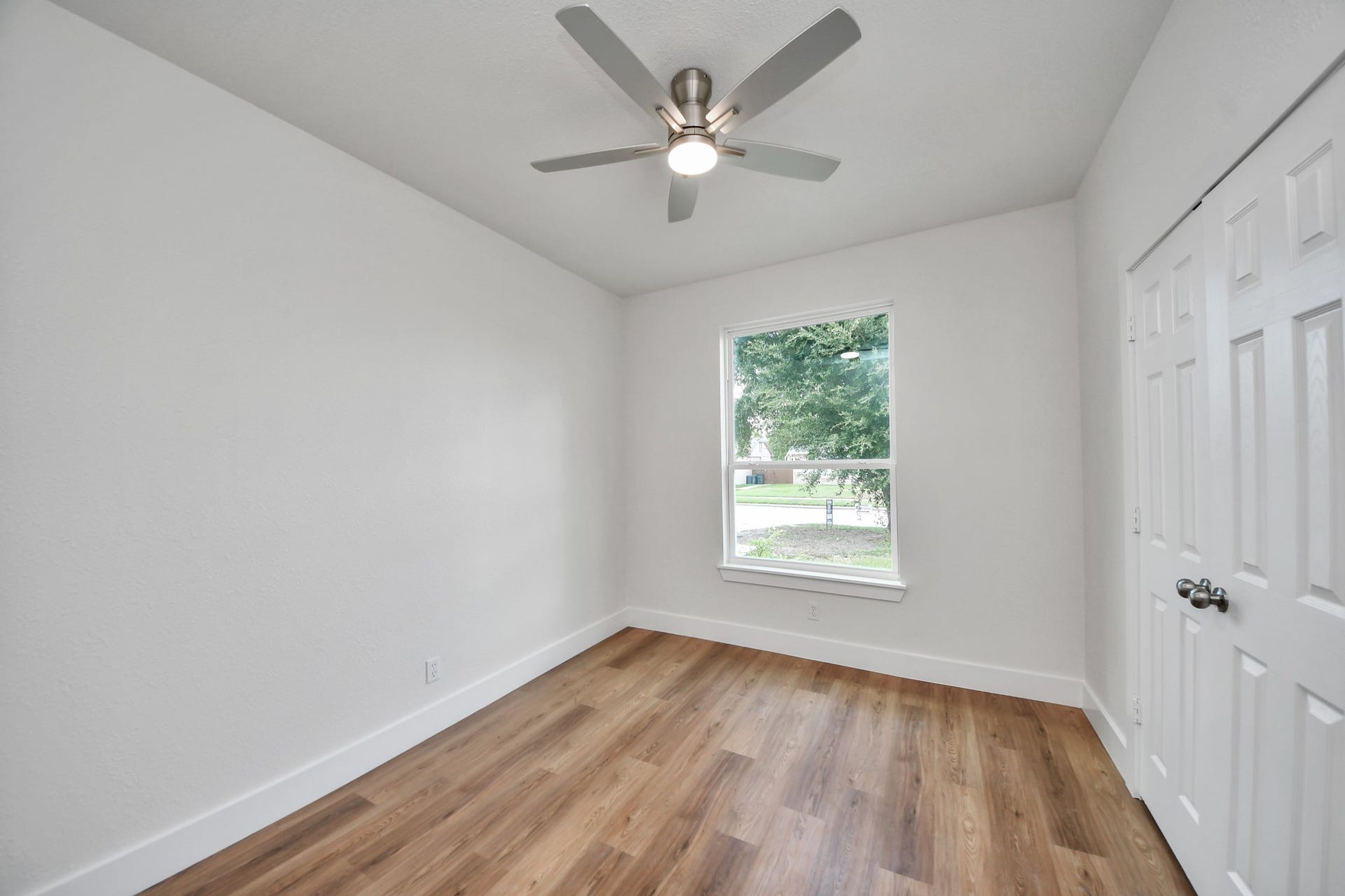 Bright and inviting room featuring warm wood flooring, a ceiling fan, and a large window offering natural light and views of the lush outdoors.