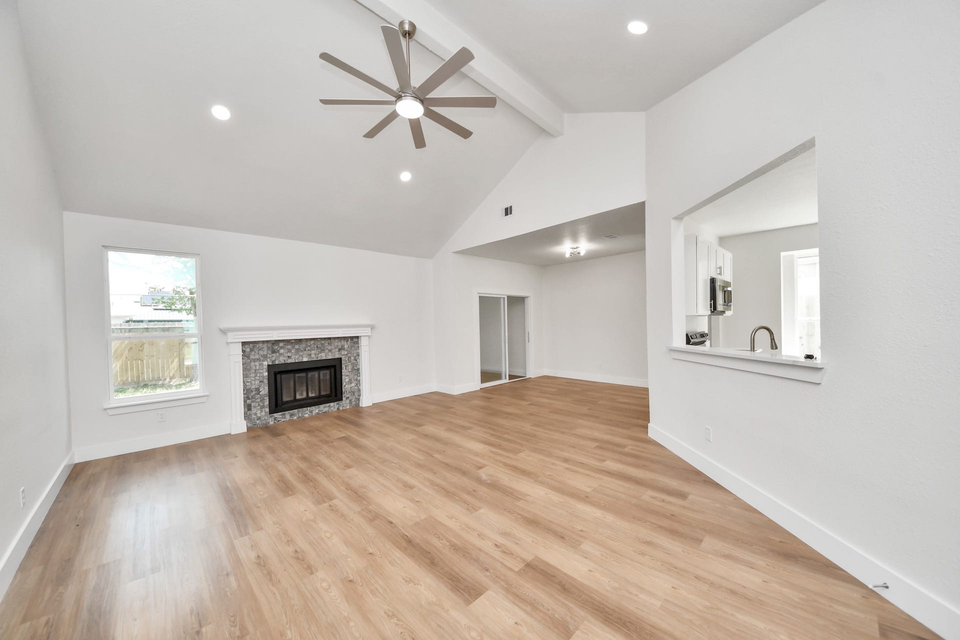 Spacious living area with vaulted ceilings and a modern ceiling fan, featuring a cozy fireplace and wood-look flooring, seamlessly connected to the kitchen through a convenient pass-through.