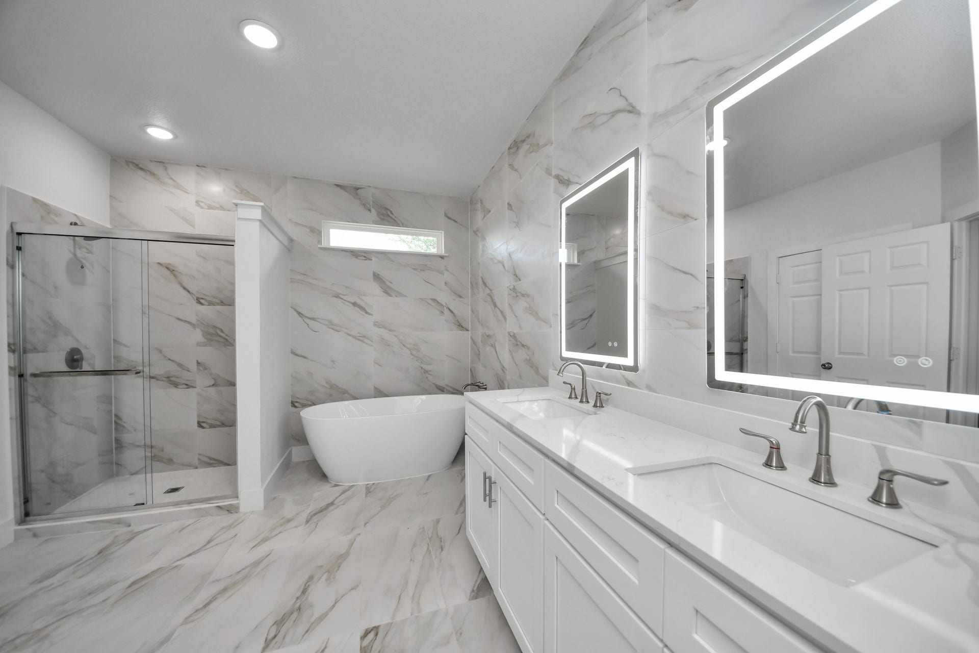 Luxurious bathroom featuring a modern freestanding tub, sleek walk-in shower, and dual vanities with illuminated mirrors, all set against elegant marble-patterned walls.