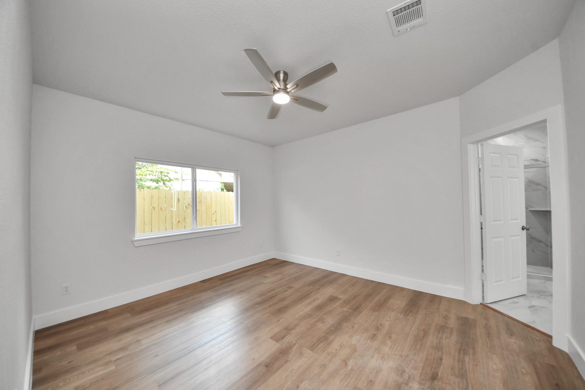 Spacious bedroom with sleek hardwood floors, a modern ceiling fan, and ample natural light from a large window; features an en-suite bathroom with elegant marble accents.