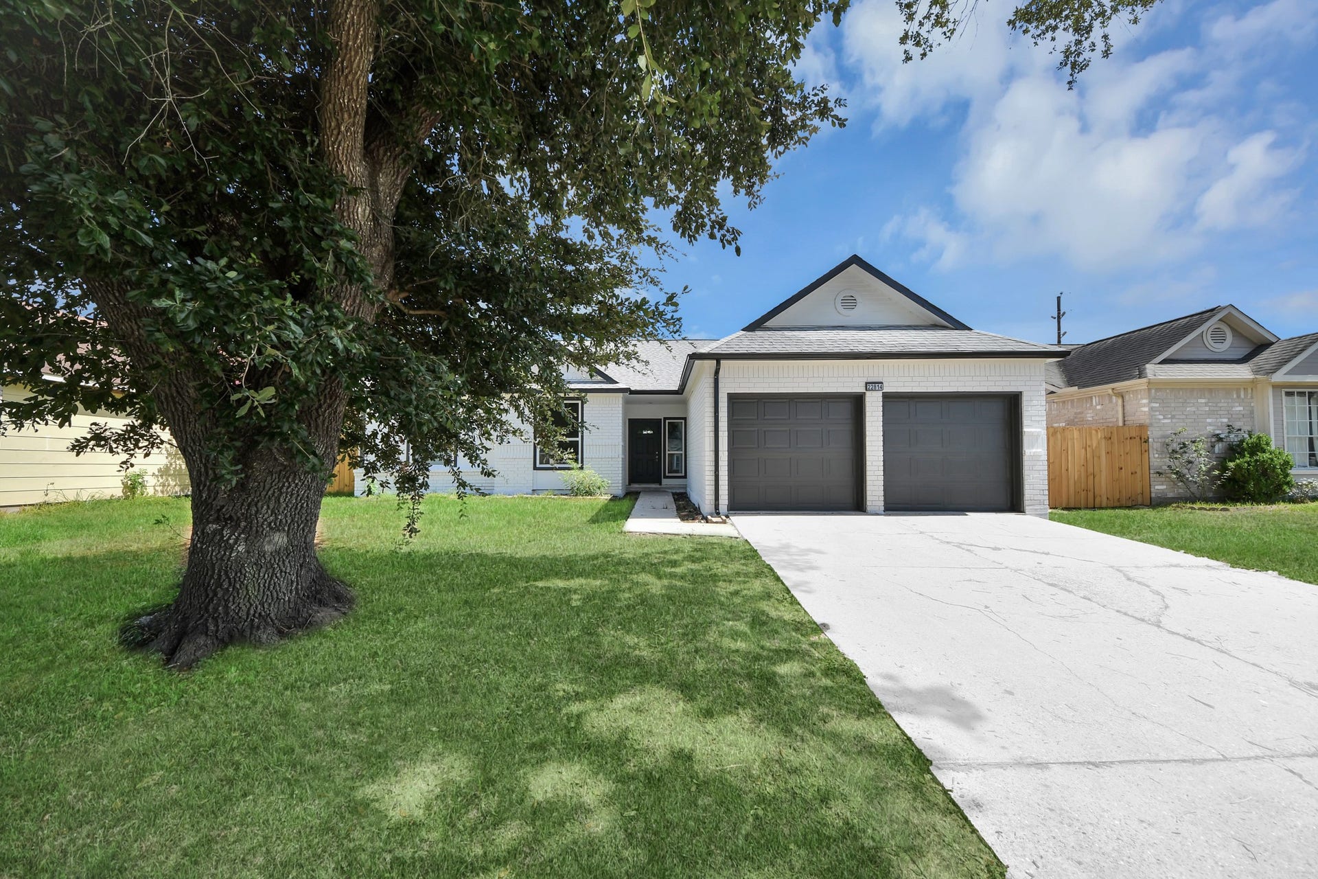 Charming home with a spacious two-car garage, nestled under a large, shady tree, offering a welcoming front yard and a long driveway for ample parking.