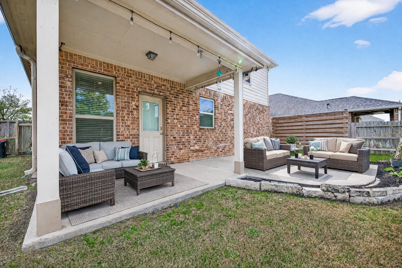 23010 Tindarey Falls Lane, Katy, TX 77493 This photo showcases a cozy backyard patio with comfortable outdoor seating on a concrete slab, set against a brick exterior. The area is partially covered, featuring string lights and a small garden space bordered by stone edging, perfect for outdoor relaxation and entertaining. (enhanced photo)