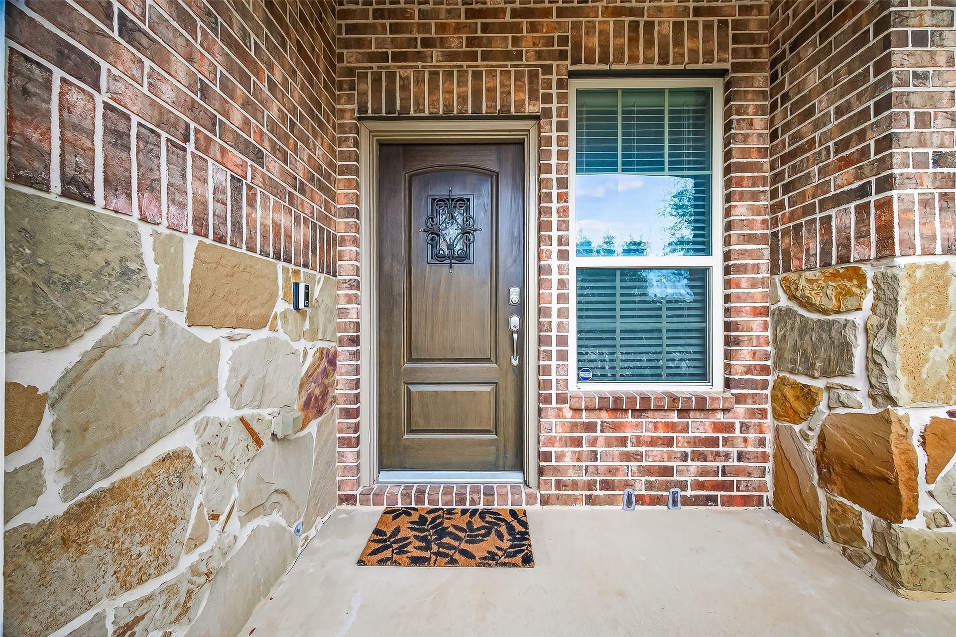 23010 Tindarey Falls Lane, Katy, TX 77493 This photo showcases a welcoming entrance with a sturdy wooden door featuring decorative ironwork. It's framed by brick and stone walls, adding a rustic charm. A large window with shutters allows natural light inside, and a doormat adds a cozy touch.