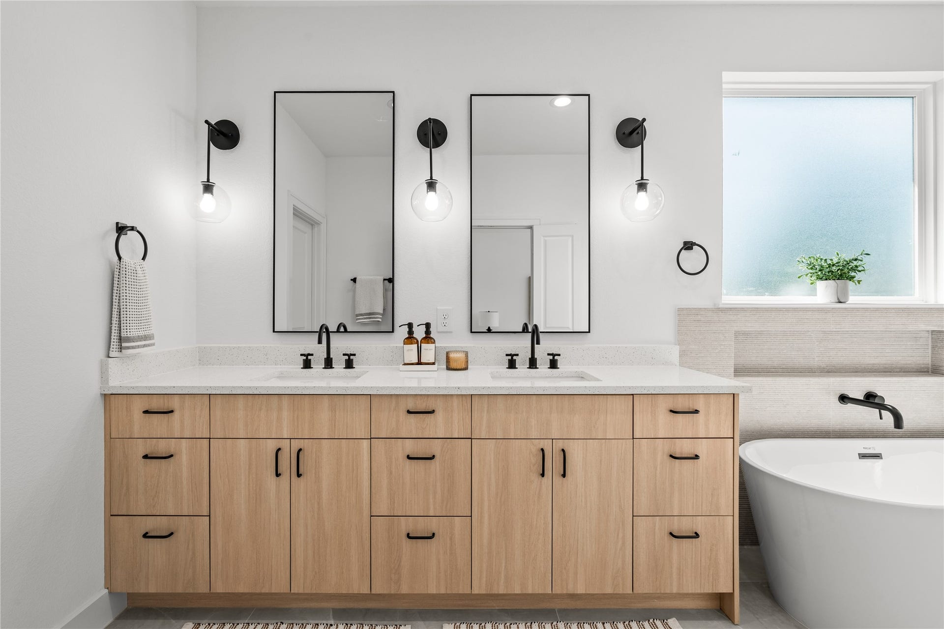 3012 & 3014 Beulah St, Houston, TX 77004 The primary bathroom features a large double vanity with wood cabinets, matte black pulls, and a white quartz countertop with dual under-mount sinks. Two black-framed mirrors are flanked by globe sconces, and a deep freestanding soaking tub sits right under a frosted privacy window.