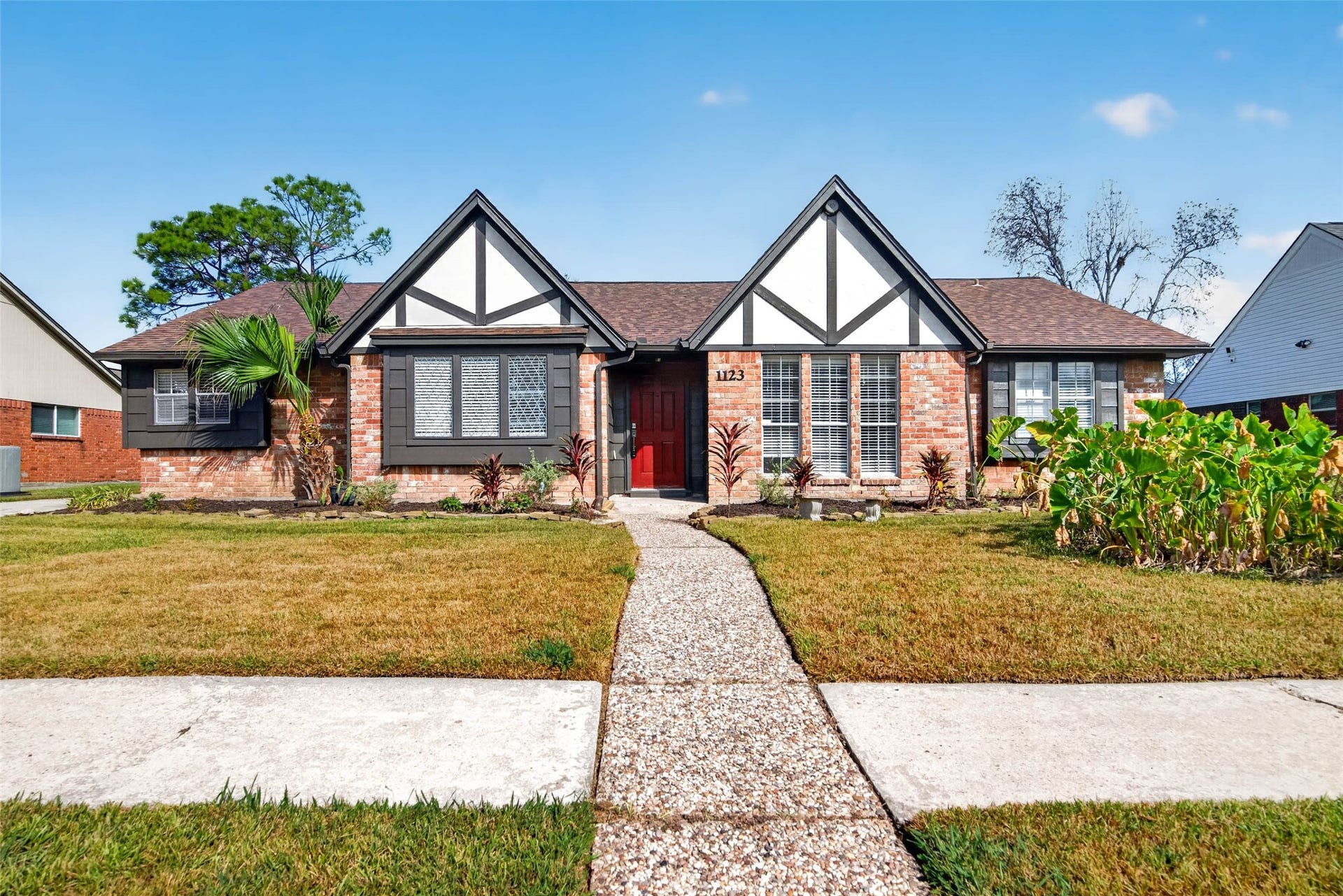 1123 Woodhorn Drive, Houston, TX 77062 Front view of the single-story ranch, with brick walls, white gables with dark timber trim, a brown shingle roof, and a red front door reached by a pebbled concrete walk.