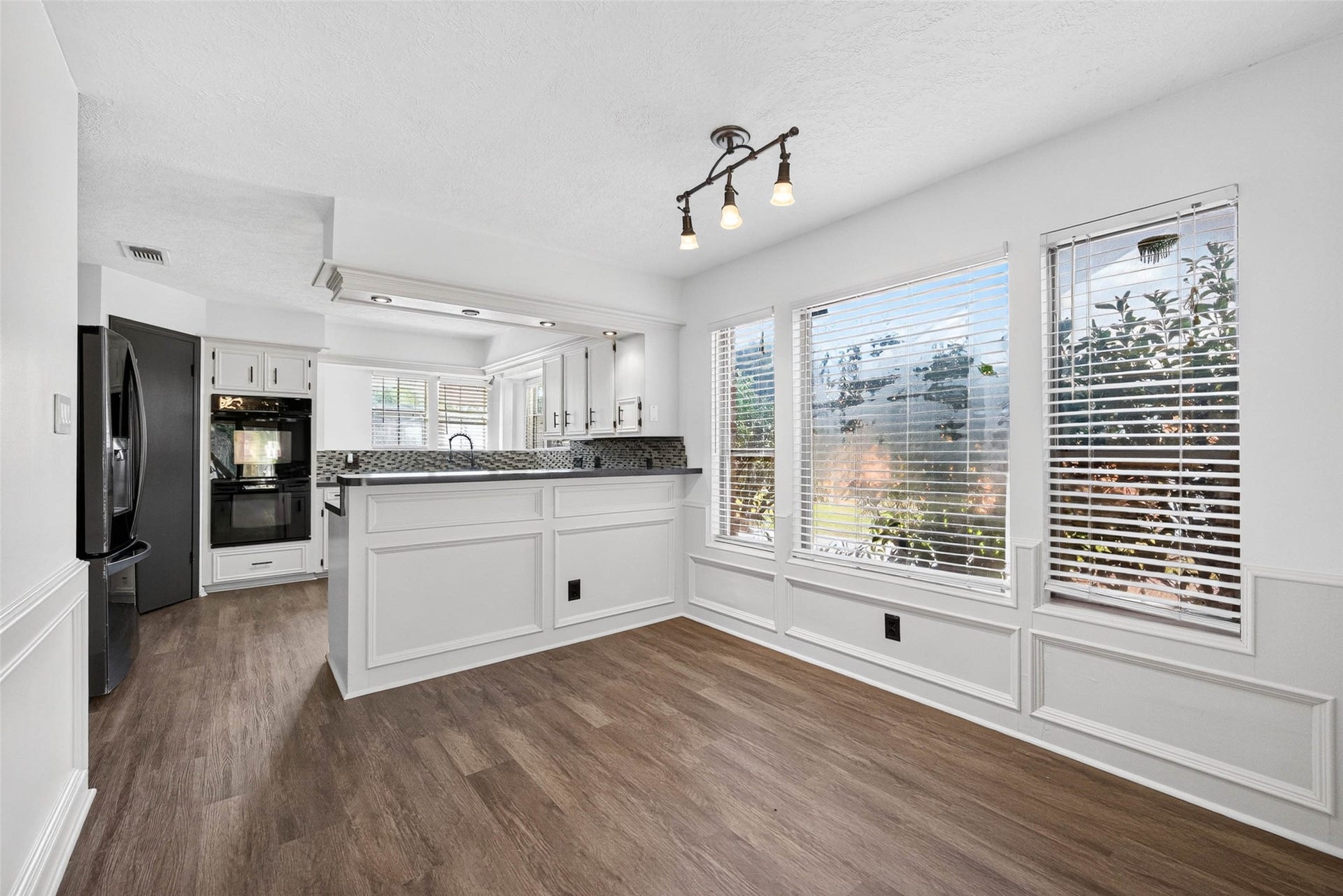 1123 Woodhorn Drive, Houston, TX 77062 This dining and kitchen space features a paneled peninsula, a mosaic tile backsplash, and a built-in black double wall oven. Large windows with blinds let in natural light, framing views over the wood-look flooring.