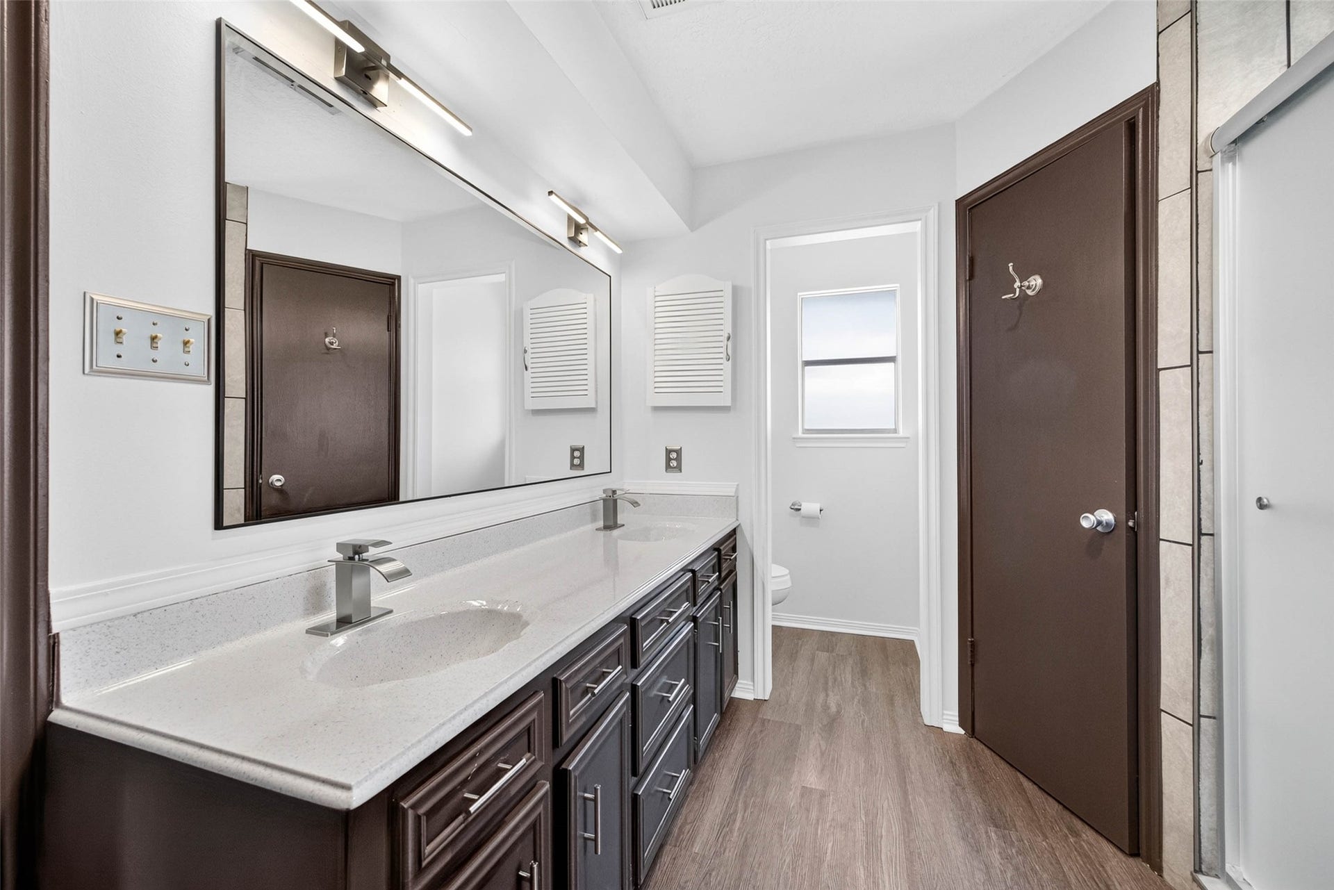 1123 Woodhorn Drive, Houston, TX 77062 En-suite bathroom with a long double vanity, large mirror, modern lighting, and a separate toilet room with a window, all on wood-look flooring.