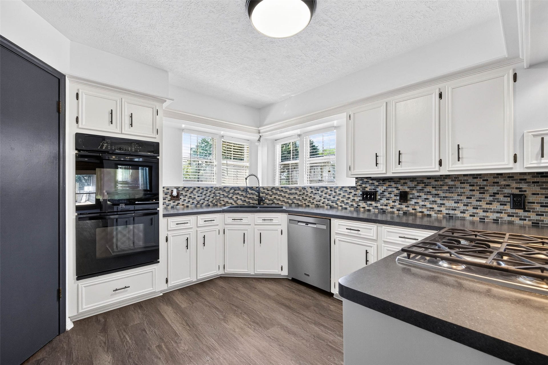 1123 Woodhorn Drive, Houston, TX 77062 Kitchen angle showing cabinets, a mosaic tile backsplash, black double wall oven, corner sink beneath windows, and a gas cooktop on the peninsula.