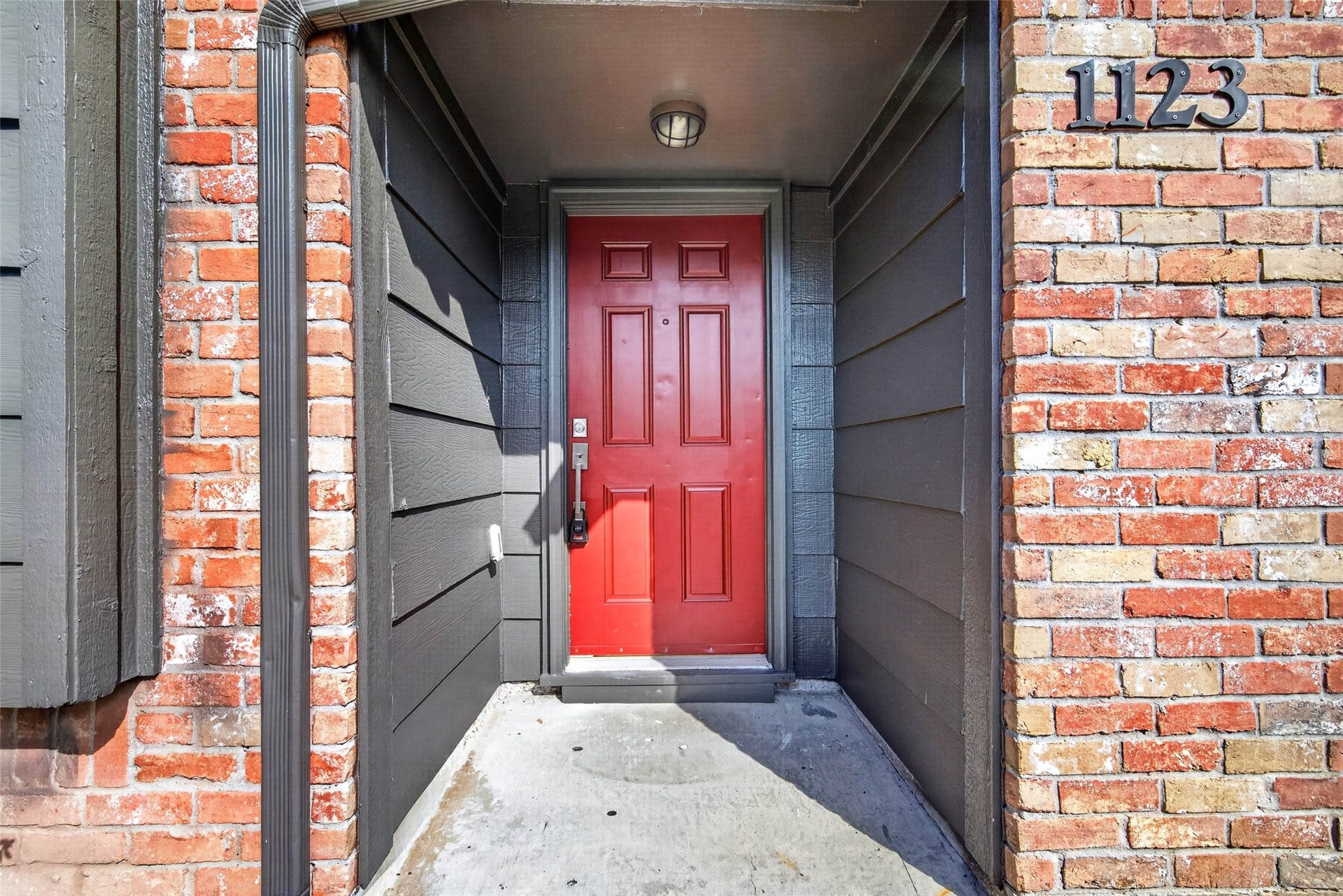 1123 Woodhorn Drive, Houston, TX 77062 Recessed front entry with a red paneled door, dark gray siding walls, and the house number 1123 in dark metal on the brick wall.