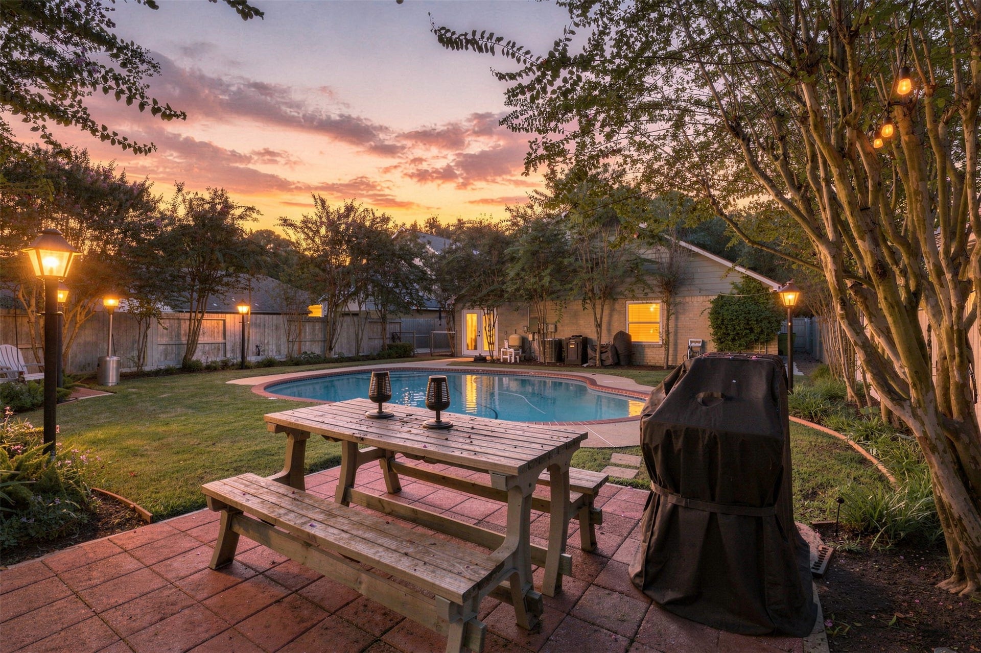 Backyard oasis at sunset showcases the sparkling pool surrounded by mature trees, landscape lighting, and an in ground gas-line-connected grill at the patio ready for entertaining under the evening sky.