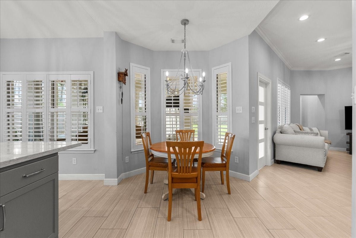 This inviting dining area features a cozy round wooden table with four chairs, elegant light gray walls, and abundant natural light through multiple windows with shutters. The open layout flows seamlessly into a comfortable living space, enhanced by warm wood-style flooring and a modern chandelier.