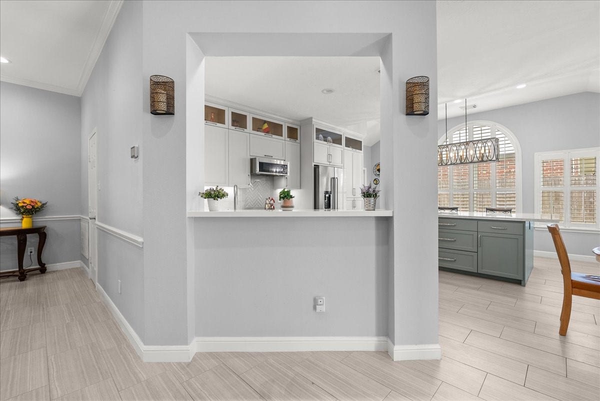 This photo showcases a modern, open-concept kitchen and dining area. It features light gray walls, sleek white cabinets, and a large kitchen island with a view into a bright dining space, highlighted by a stylish light fixture and arched window. The area is spacious and inviting.