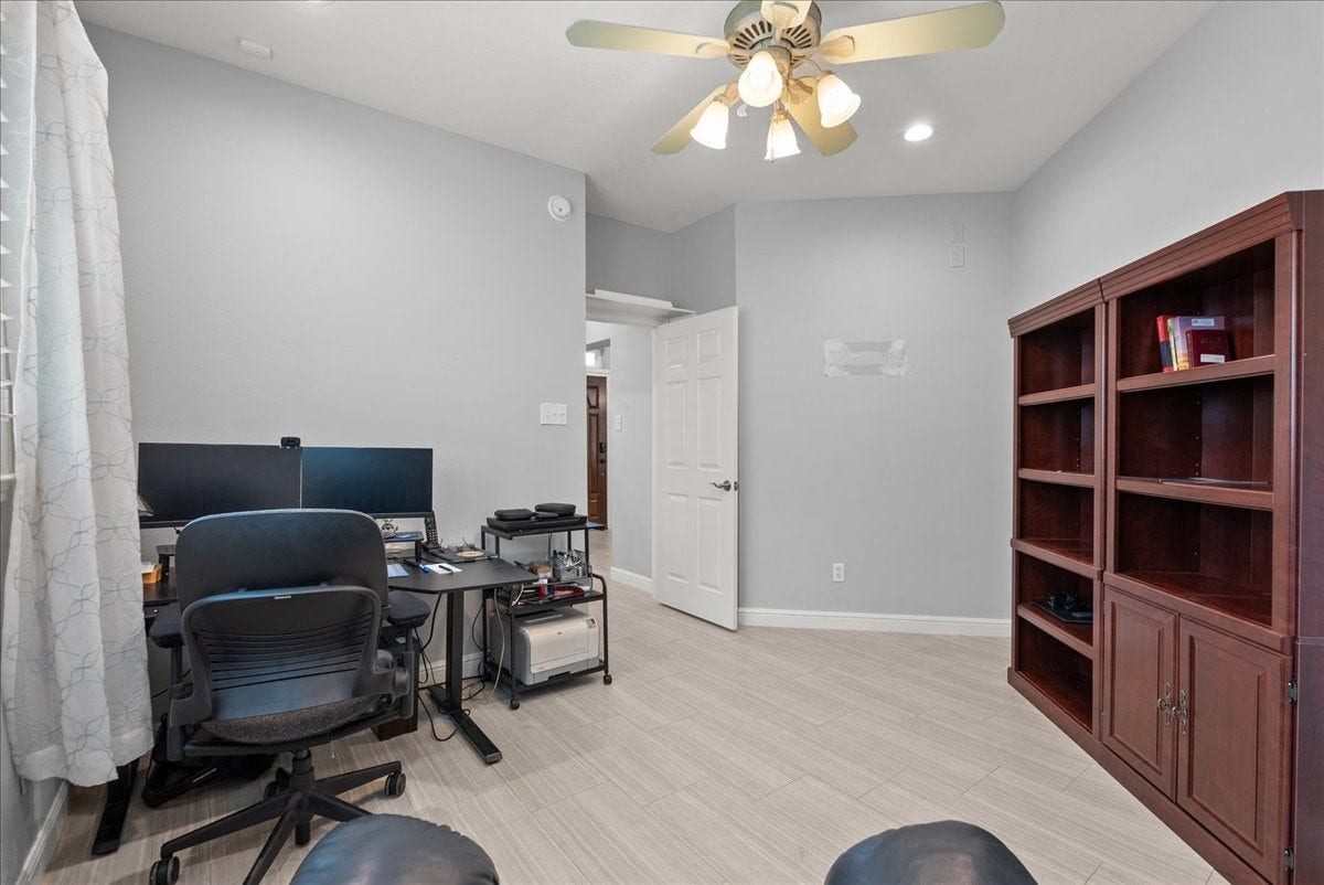 This room features a modern home office setup with a desk, dual monitors, and a comfortable chair. It has light gray walls, a ceiling fan with lights, and ample natural light from a window. A large wooden bookcase provides additional storage space. Perfect for remote work or study.
