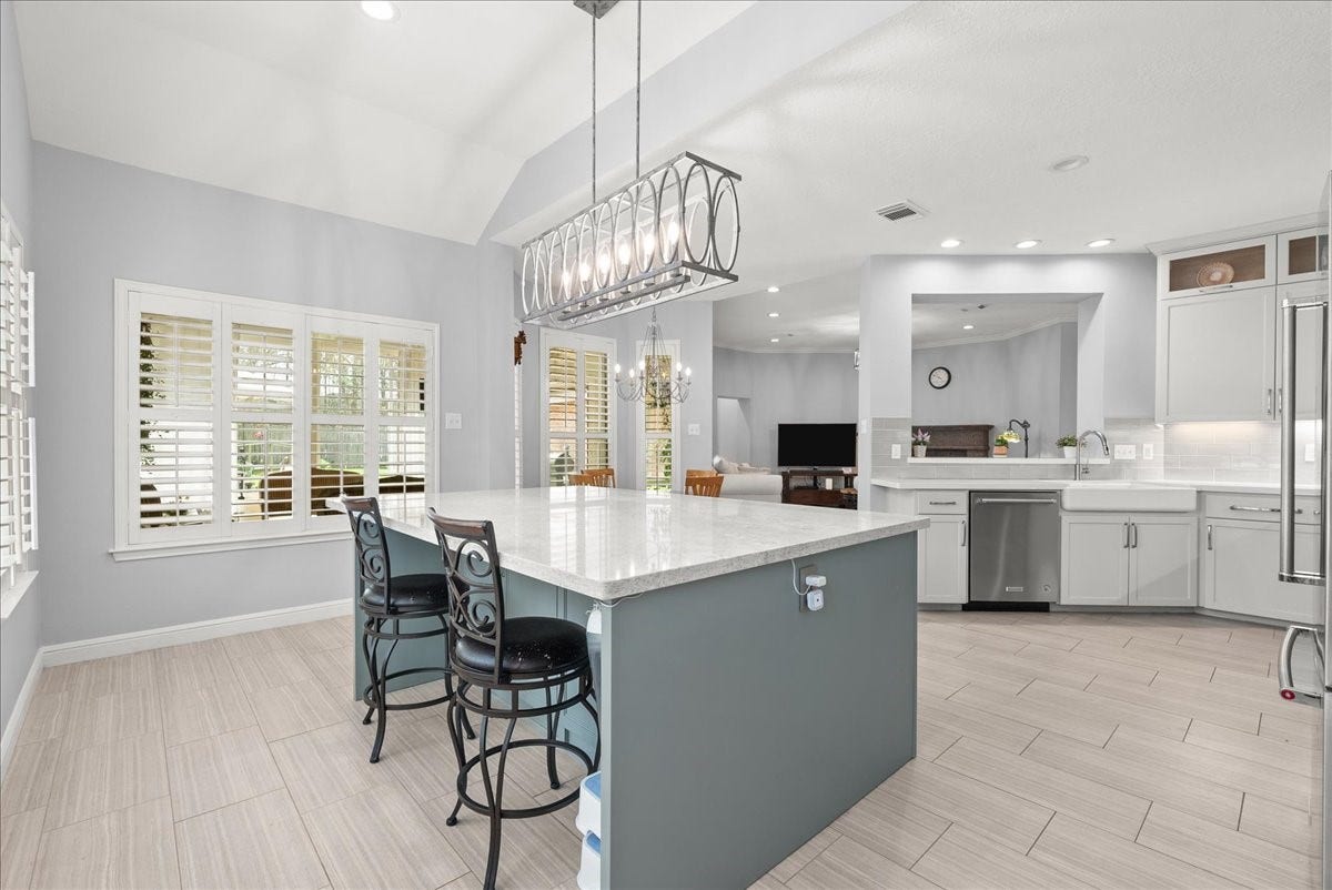 This photo showcases a modern, open-concept kitchen with a spacious island featuring a quartz countertop and stylish pendant lighting. The kitchen includes stainless steel appliances, ample cabinetry, and a bright dining area with large windows and plantation shutters. The space flows seamlessly into a cozy living area, offering a perfect layout for entertaining.
