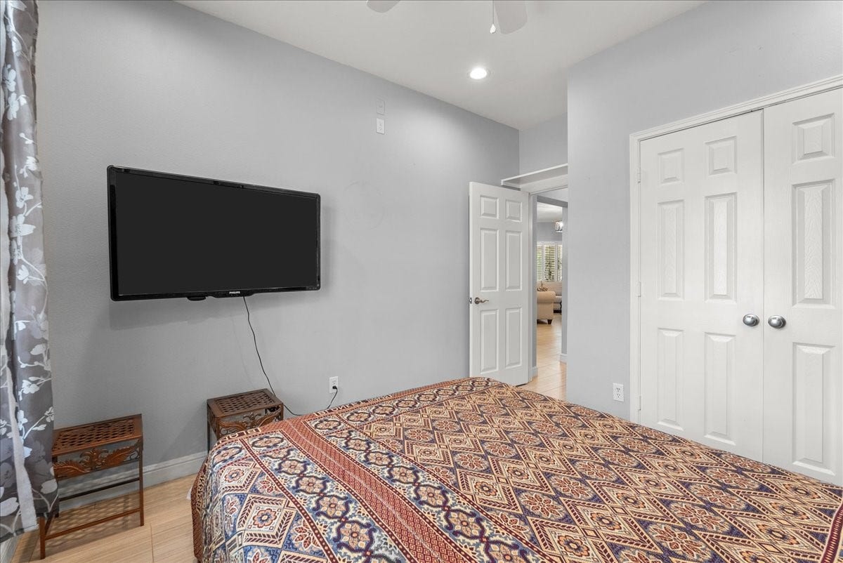 This cozy bedroom features light gray walls, a mounted flat-screen TV, and a patterned bedspread. It includes a double-door closet and an open doorway leading to the living area, creating a spacious feel.