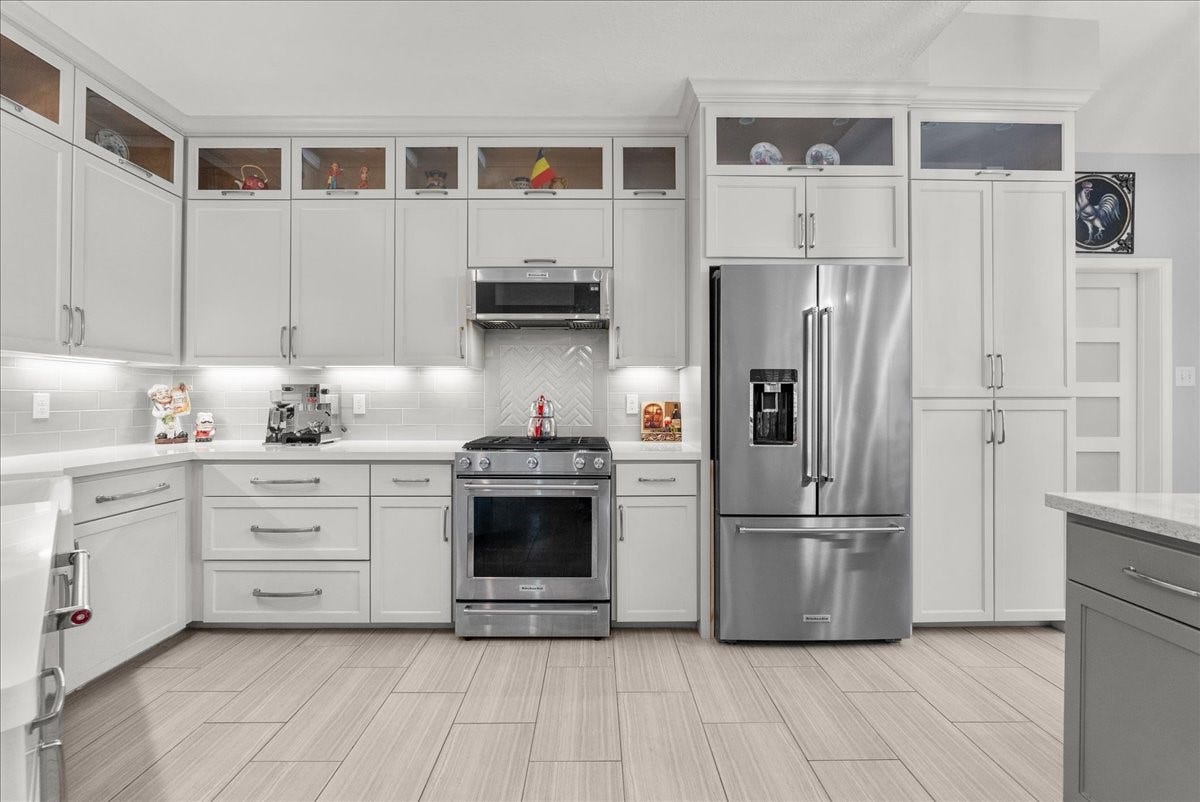 This kitchen features modern white cabinetry with glass accents, stainless steel appliances, and a sleek tile backsplash. The layout is spacious and well-lit, offering ample storage and a contemporary design.