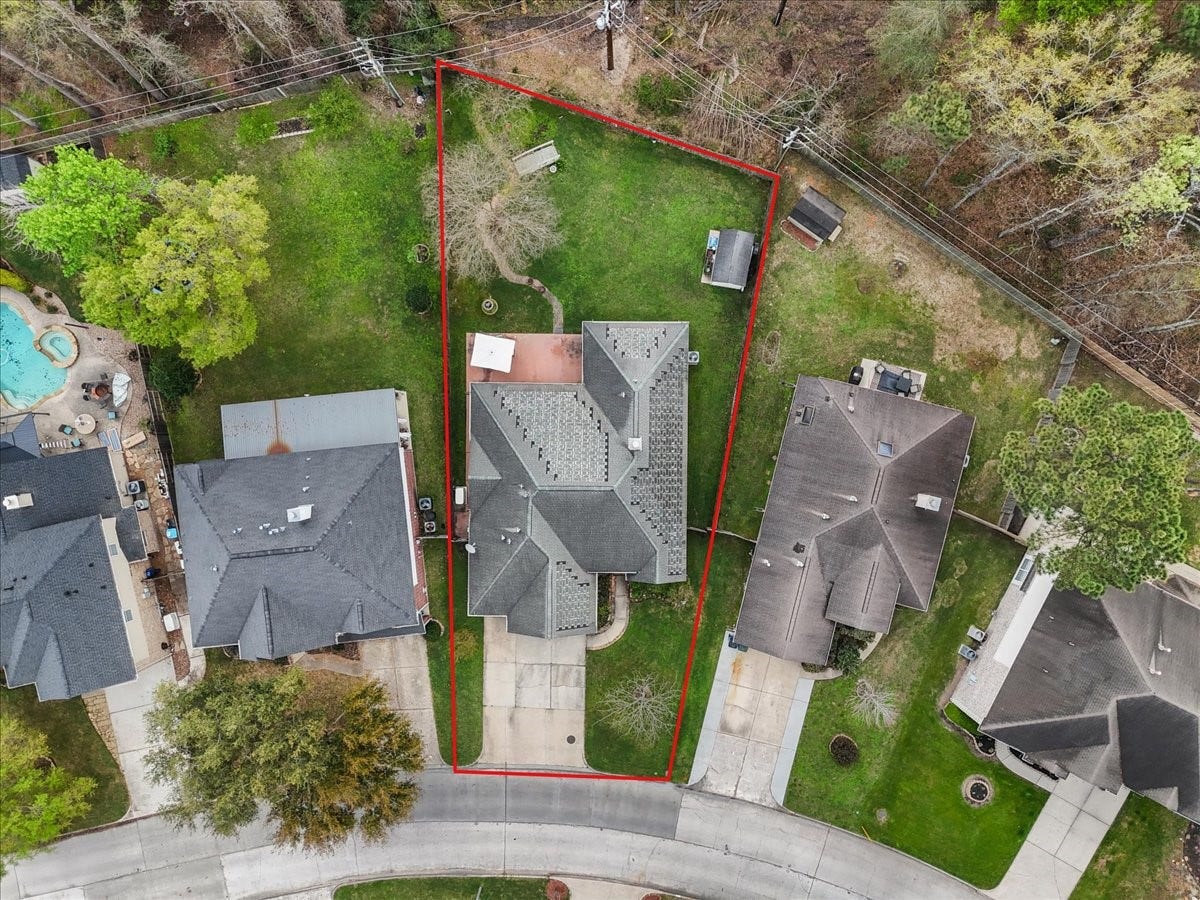 This aerial photo shows a uniquely shaped property with a spacious backyard, outlined in red. The house has a large roof and driveway, situated in a quiet suburban neighborhood with neighboring homes and greenery.