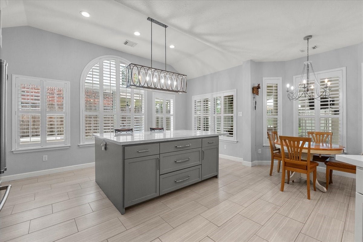 Bright, modern kitchen with large windows and shutters, featuring a central island with storage, stylish lighting, and an adjacent dining area.