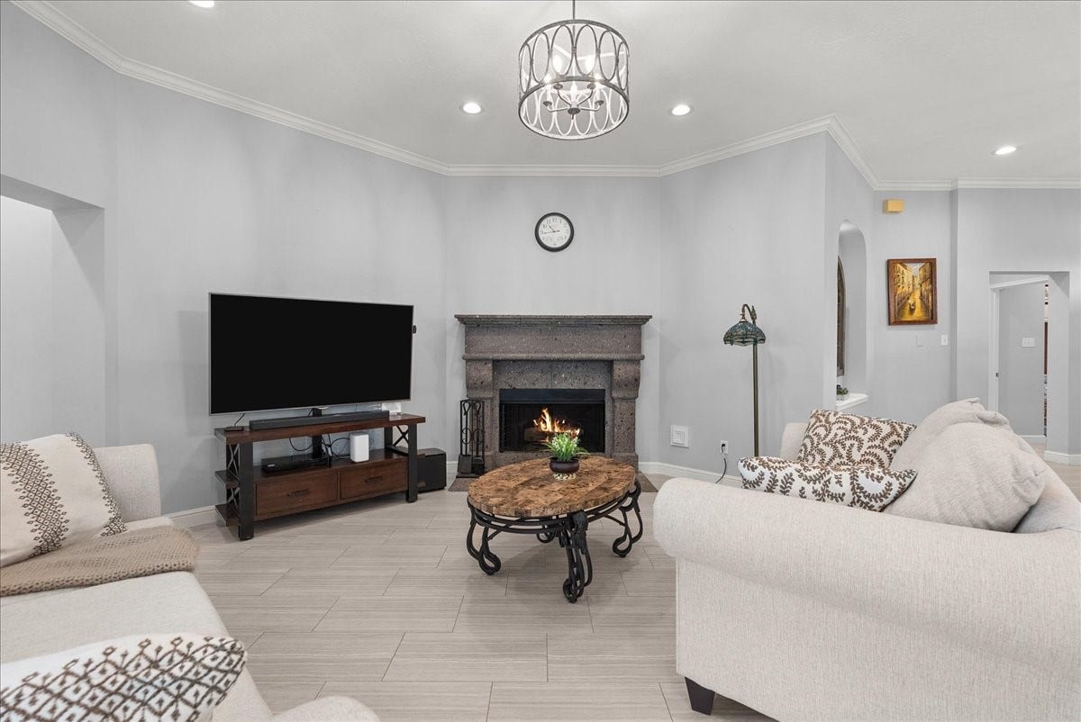 This cozy living room features a modern design with neutral tones, a stylish chandelier, and recessed lighting. It has a large TV, a decorative stone fireplace, and comfortable seating, perfect for relaxation.
