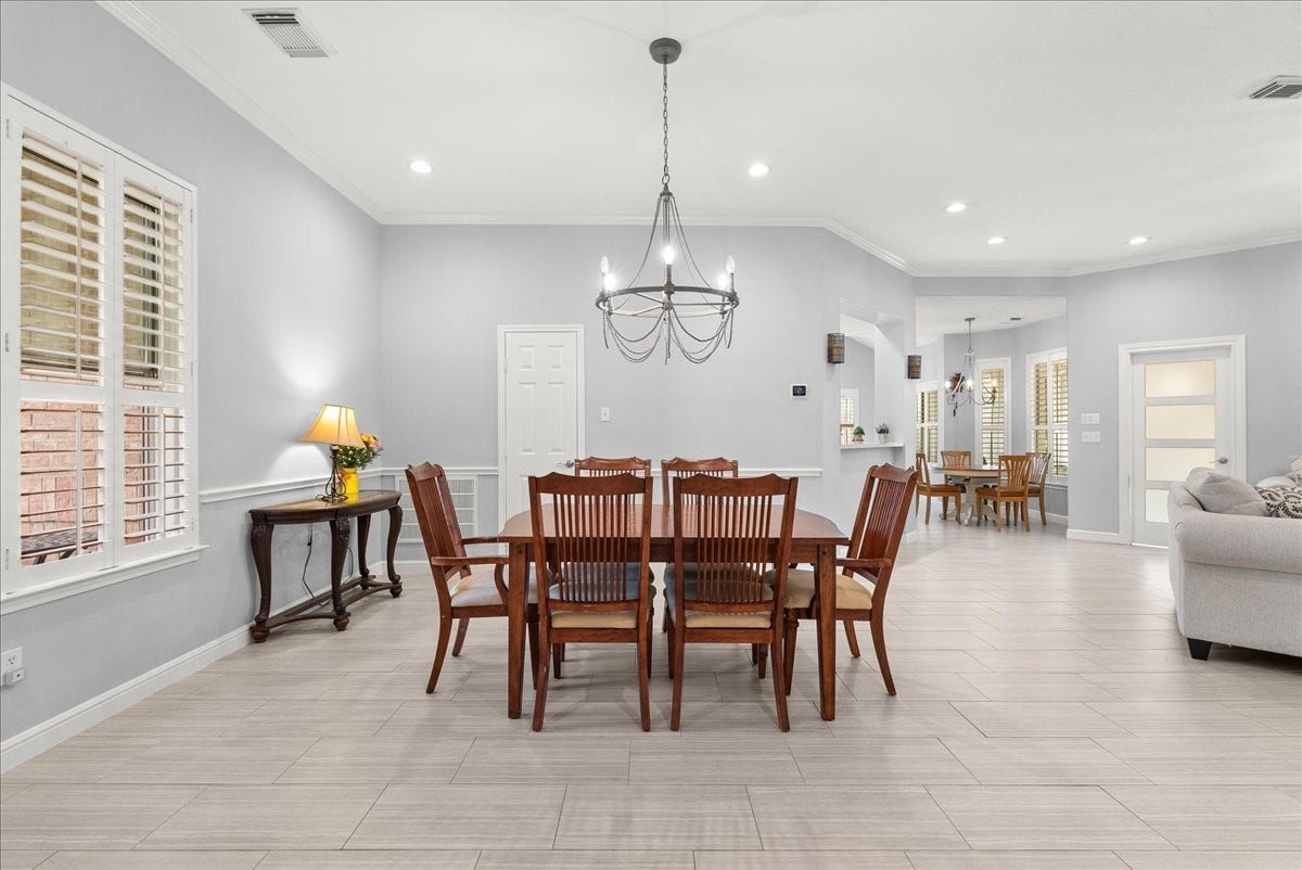 This inviting home features an open-concept dining and living area with elegant light gray walls and wood-look tile flooring. A stylish chandelier hangs above the dining table, while ample natural light fills the space through large windows with white shutters. Cozy and modern, its perfect for entertaining.