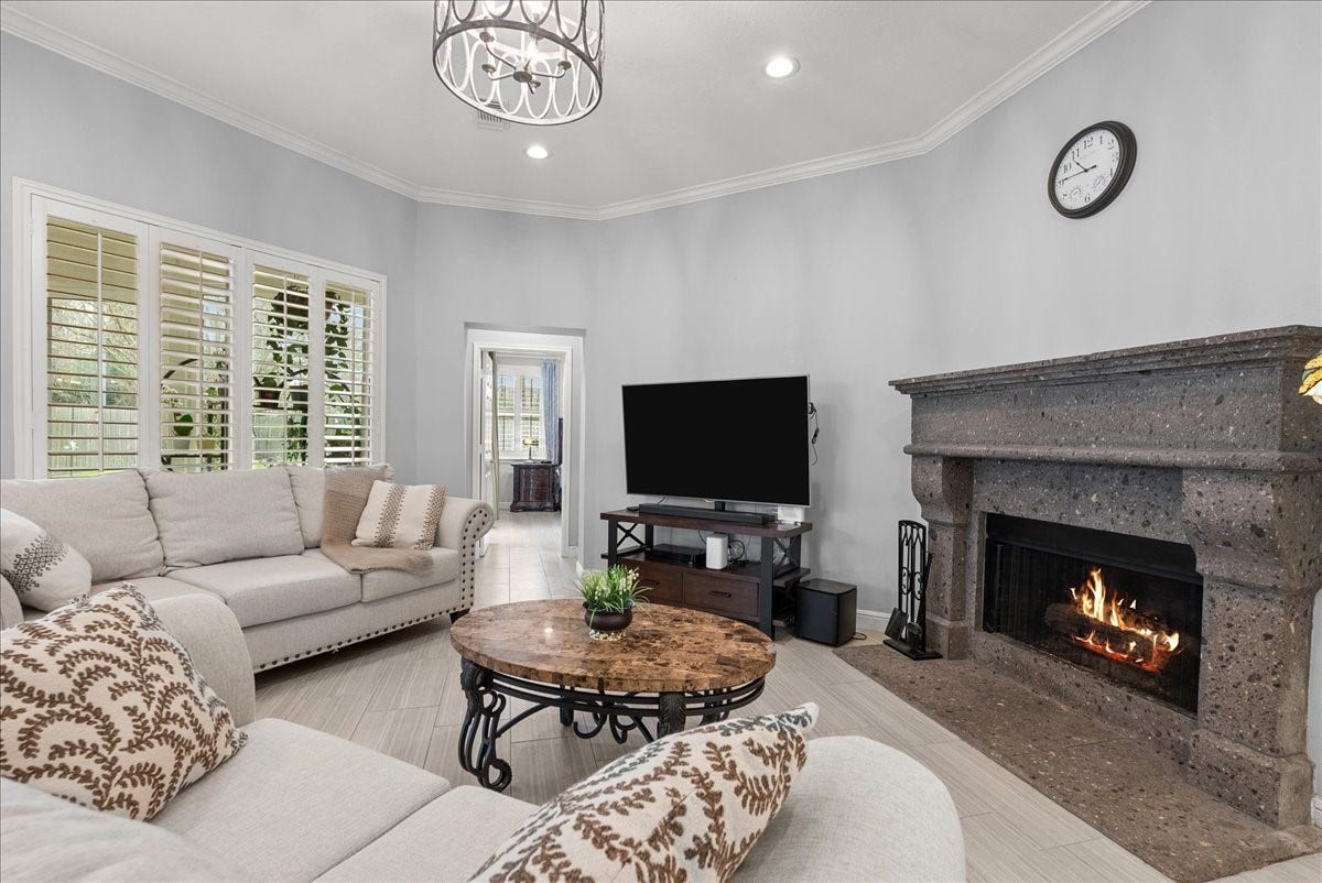 This cozy living room features a large sectional sofa, a stylish chandelier, and a warm fireplace. It has a wall-mounted TV, elegant stone coffee table, and bright windows with plantation shutters, creating an inviting and comfortable space.