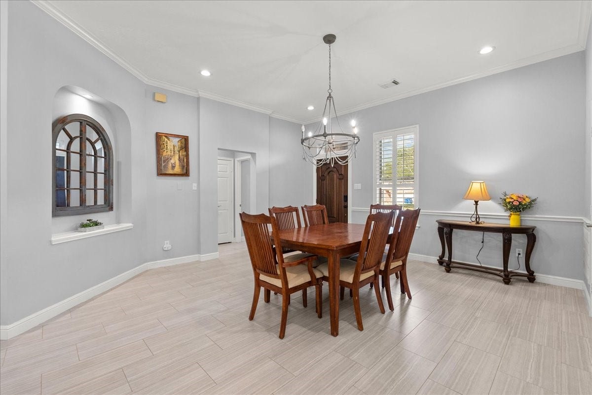 This dining room features a spacious layout with light gray walls and elegant crown molding. It includes a wooden dining table with chairs under a stylish chandelier. Natural light streams in through a window with shutters, while a decorative arched window and wall art add charm. A side table with a lamp and flowers completes the inviting atmosphere.