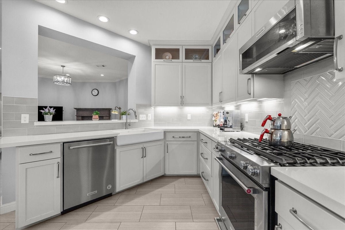 This modern kitchen features sleek white cabinetry, stainless steel appliances, and a stylish herringbone tile backsplash. It includes a gas stove, built-in microwave, and dishwasher. The large farmhouse sink and open layout offer a spacious, inviting feel, perfect for cooking and entertaining.