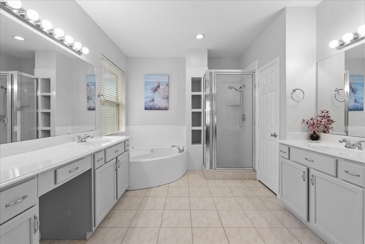 This spacious bathroom features dual vanities with ample storage, a large mirror, a corner bathtub, and a glass-enclosed shower. It is well-lit with modern fixtures and has neutral tile flooring.